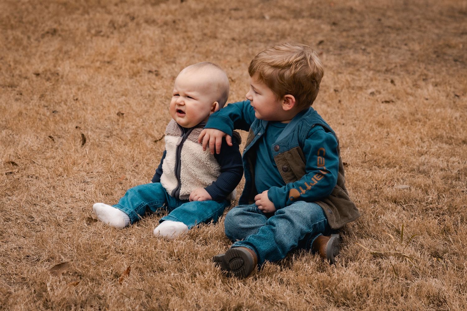 Two young children sit together on brown grass wearing coordinating blue and gray outfits.