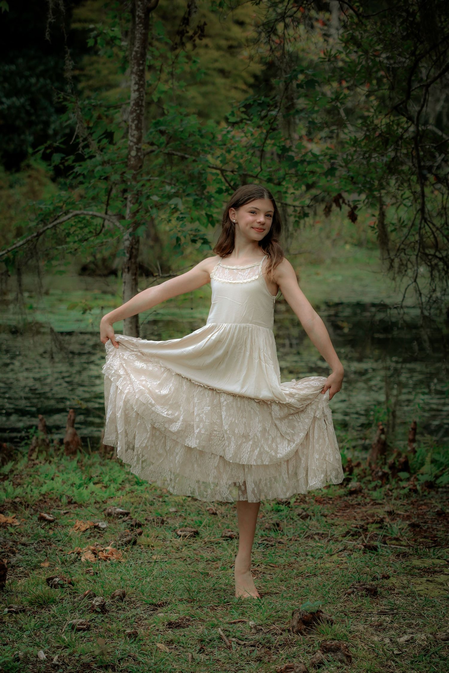 A barefoot dancer twirls in a white lace dress among pine trees.