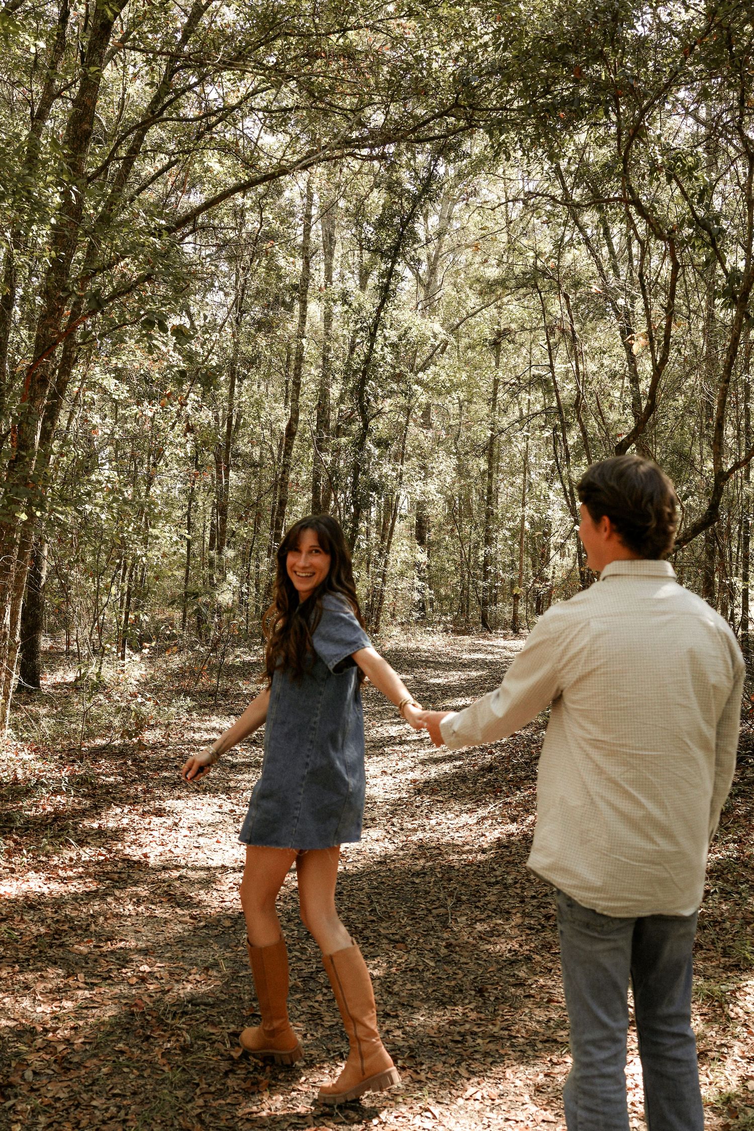 A playful couple dances and spins together in a wooded area with filtered sunlight.
