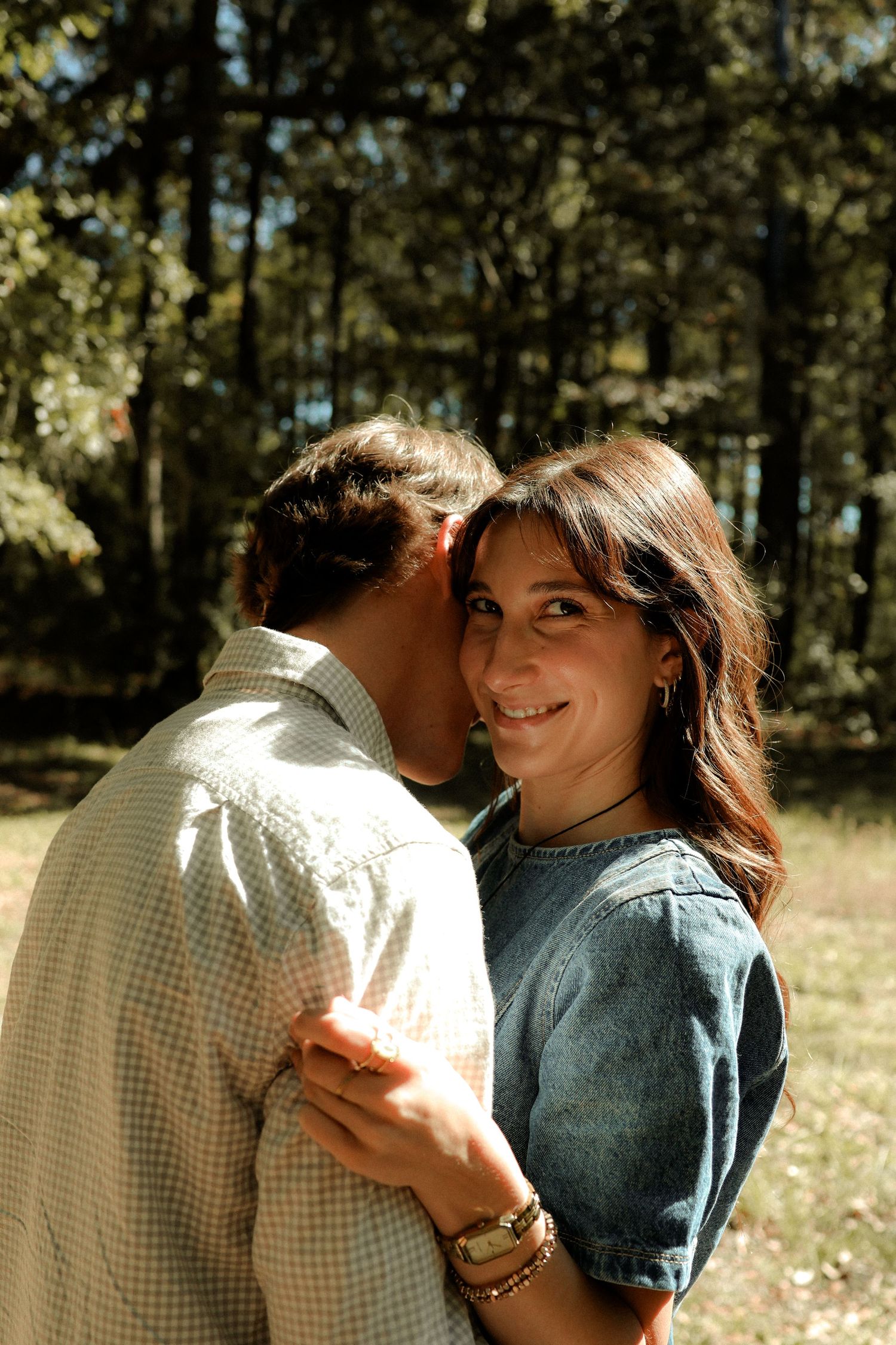 A couple shares an intimate moment in dappled sunlight among trees in a forest setting.