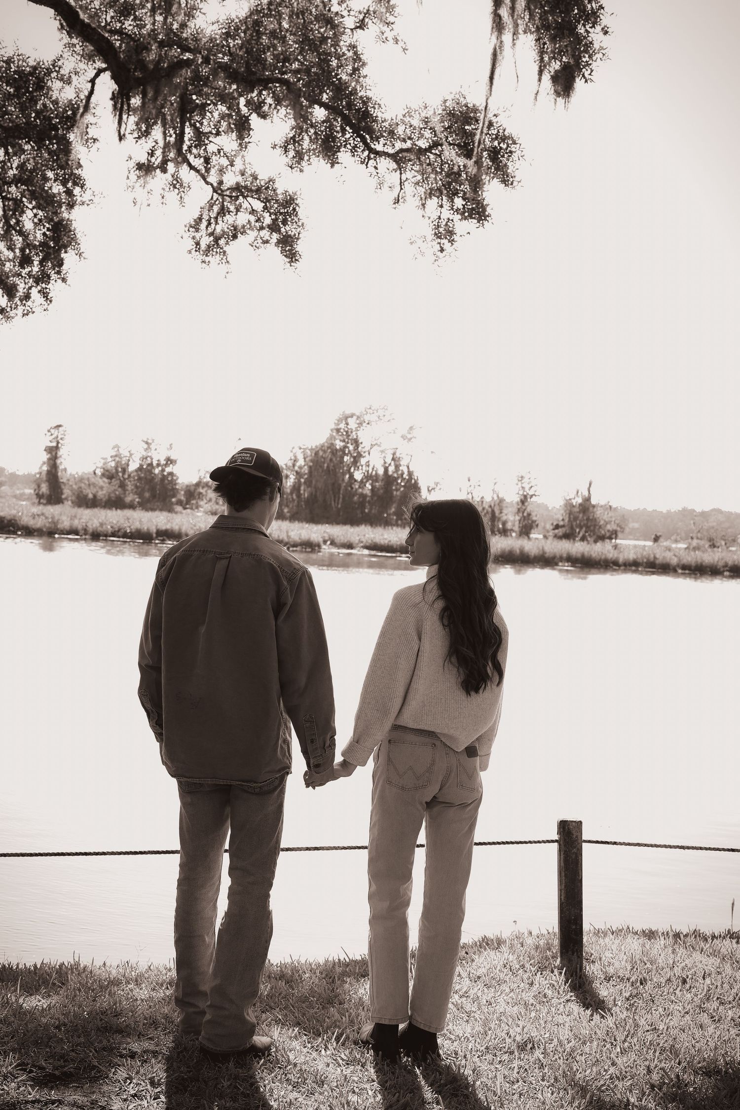 A couple stands together by a fence overlooking a serene waterfront landscape in black and white.