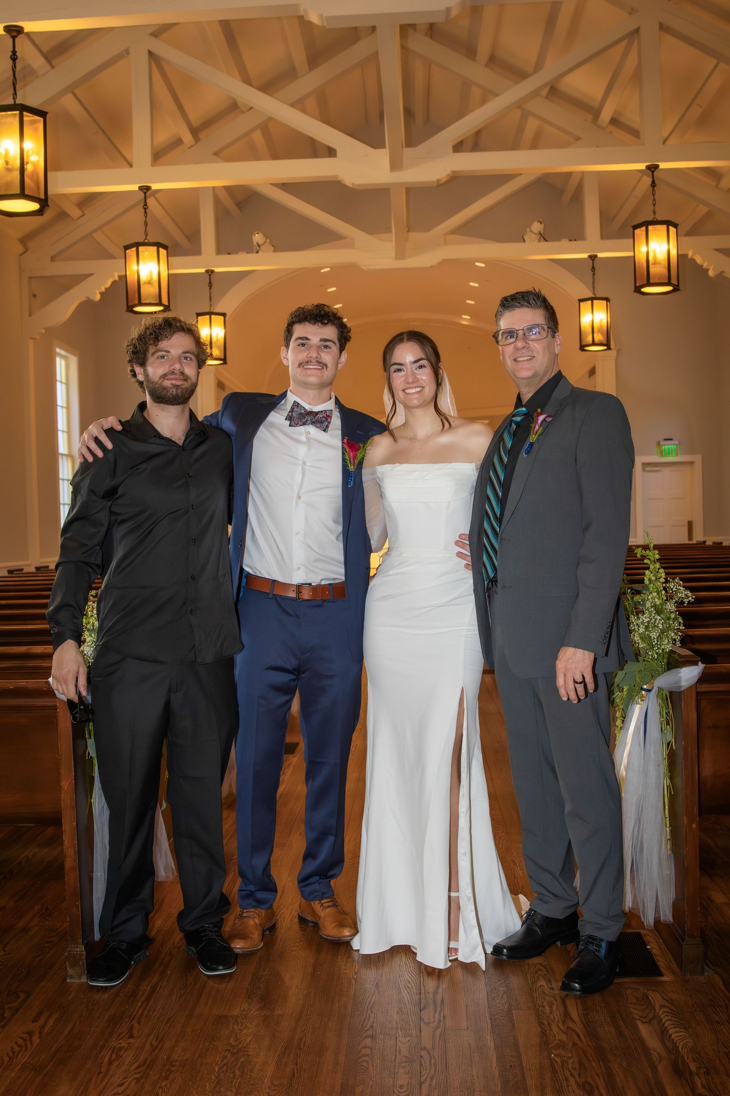 Wedding party members pose together on hardwood floor under rustic wooden beams and pendant lights.