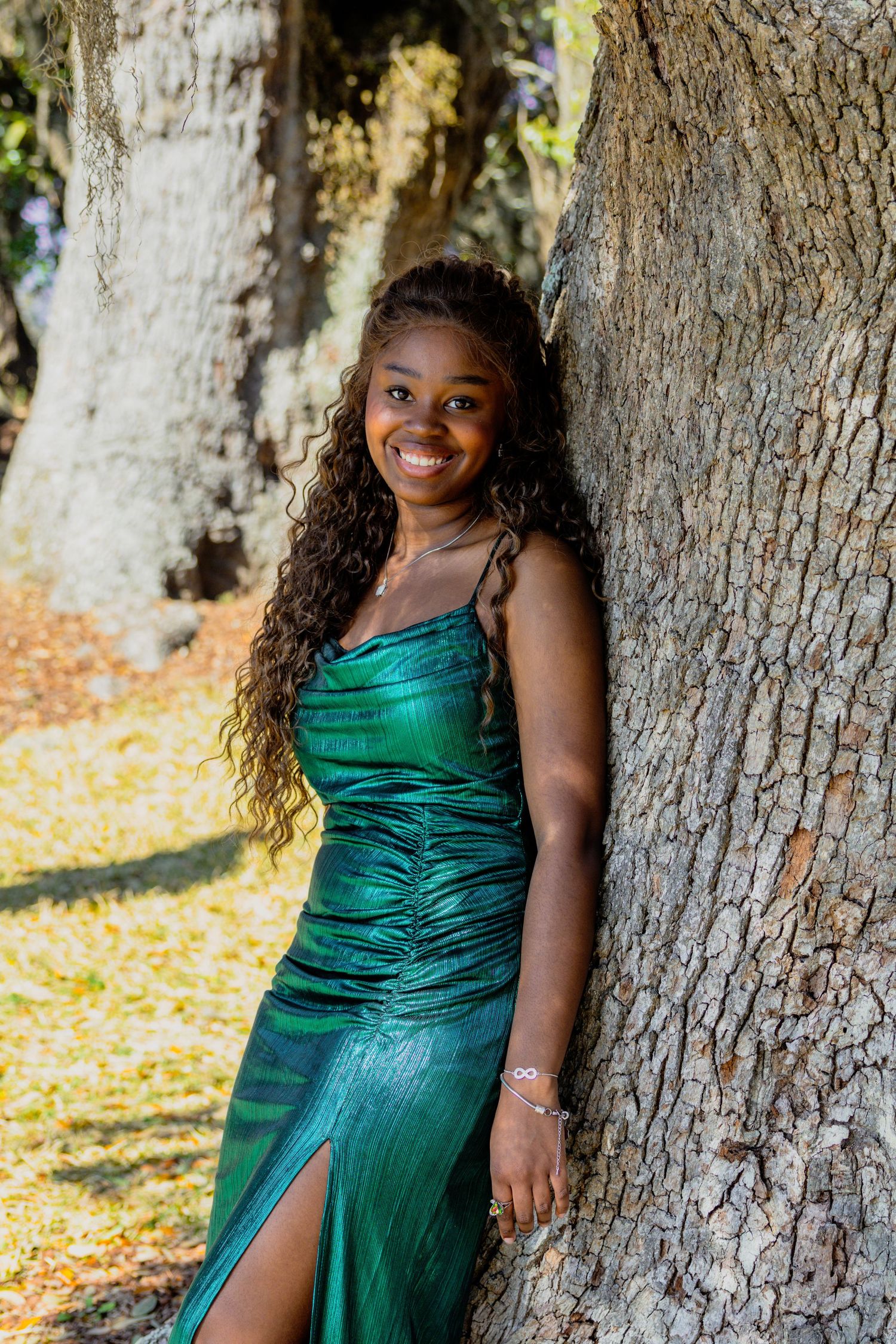 A woman in an elegant emerald green evening gown poses against a tree trunk in natural outdoor lighting.