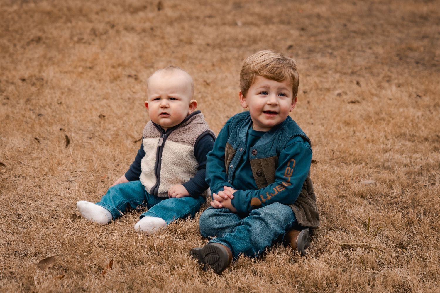 Two young children sit together on brown grass wearing coordinating blue and gray outfits.