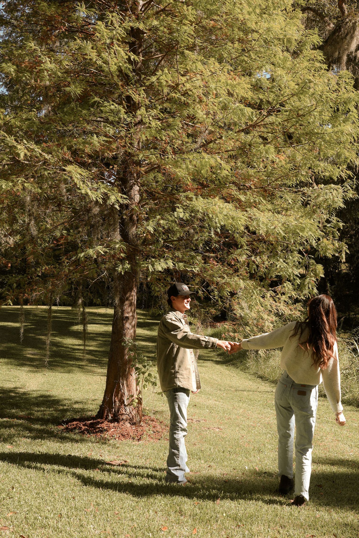 A couple holds hands and spins in a grassy park setting on a sunny afternoon.