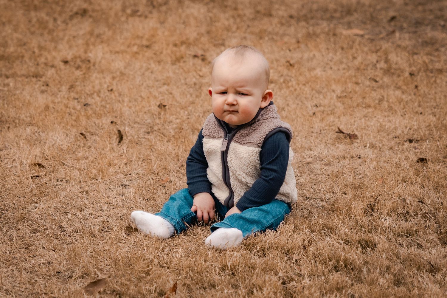 Two young children sit together on brown grass wearing coordinating blue and gray outfits.