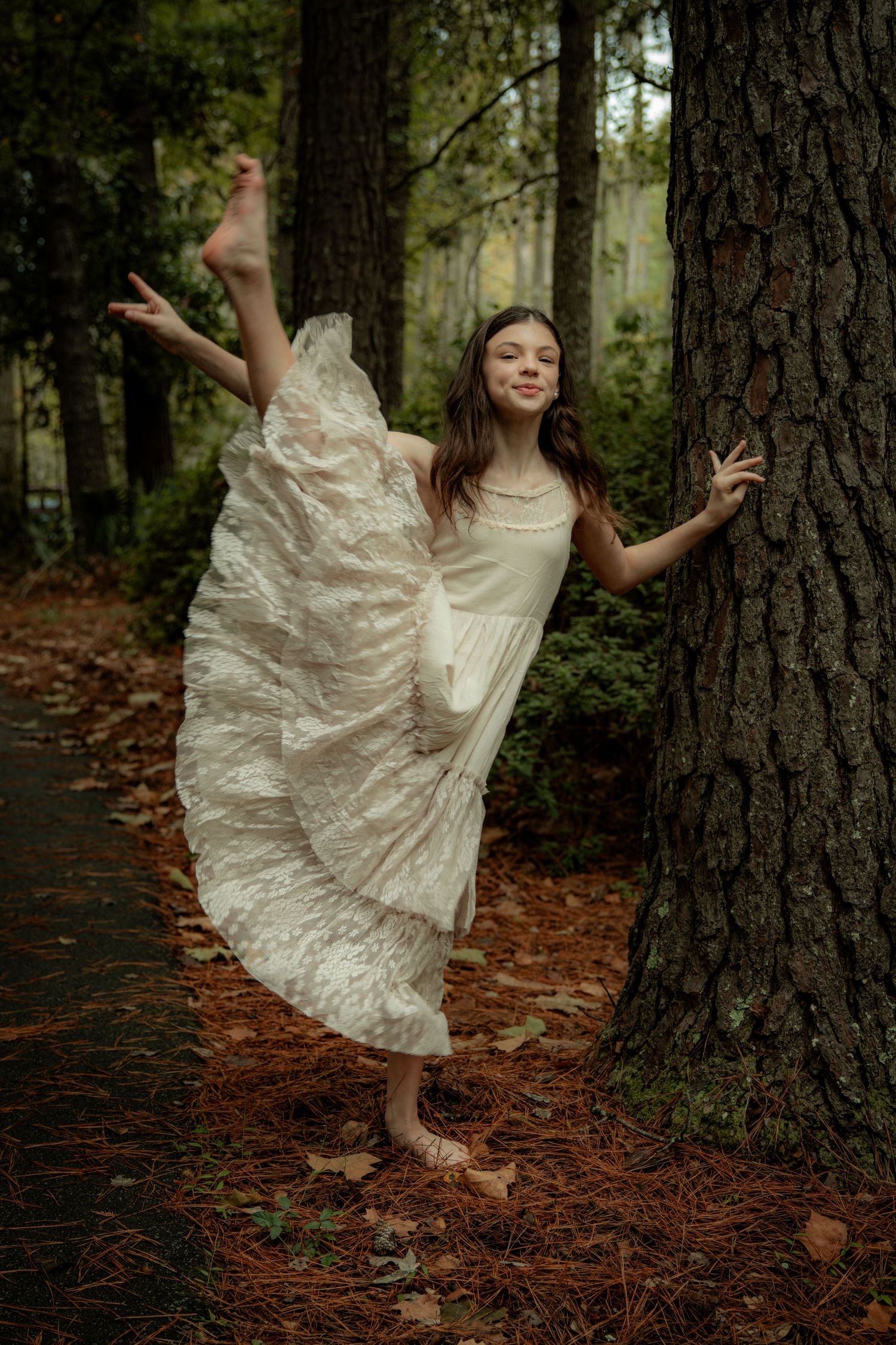 A dancer performs a playful pose against a tree trunk wearing a flowing cream-colored dress.