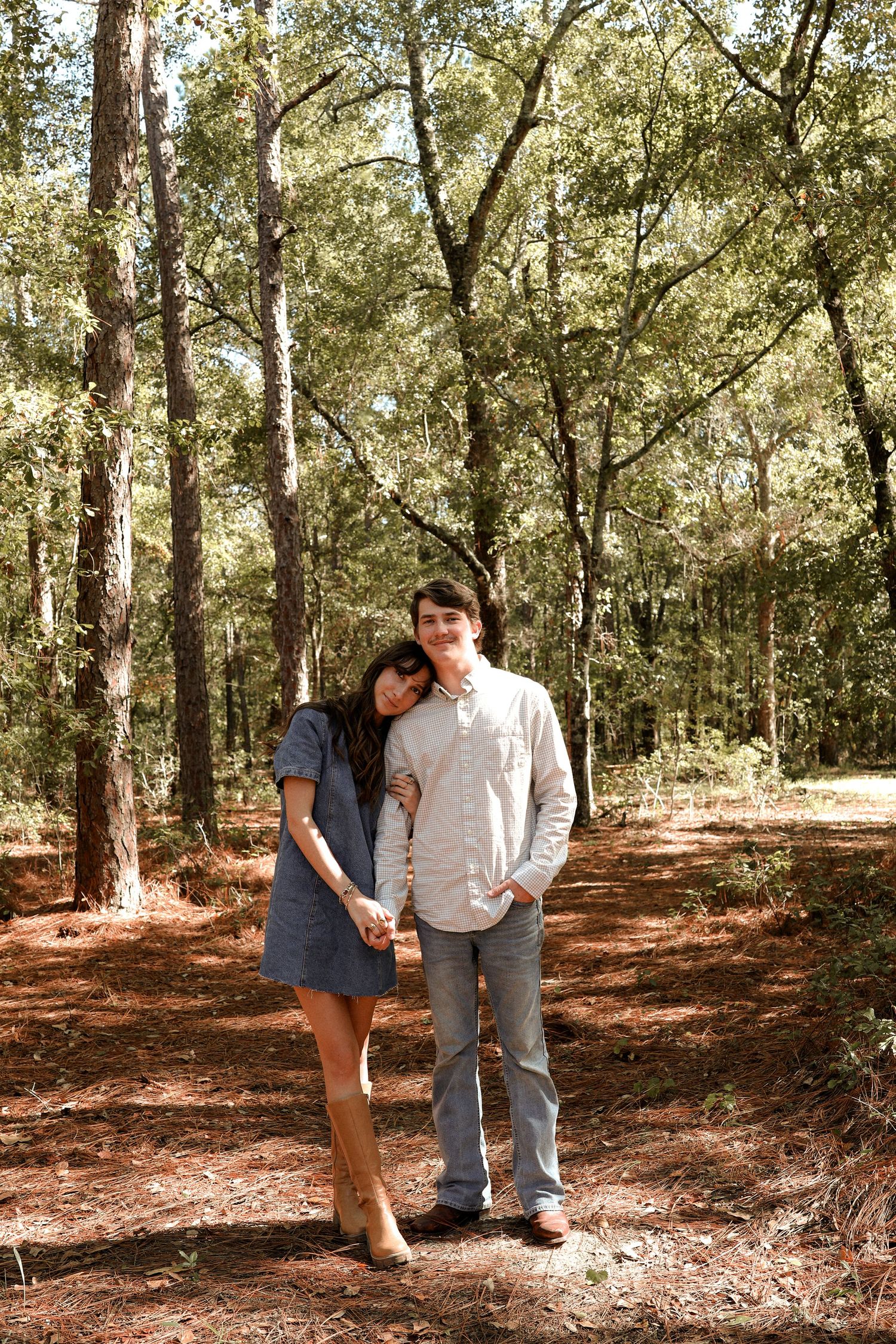A couple shares a playful moment together in a sunlit wooded area covered in fallen leaves.