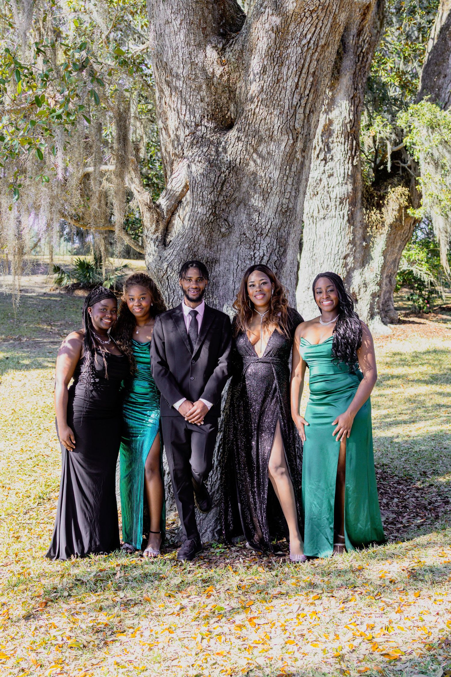 Wedding group photo beneath large oak trees with autumn leaves scattered on the ground.