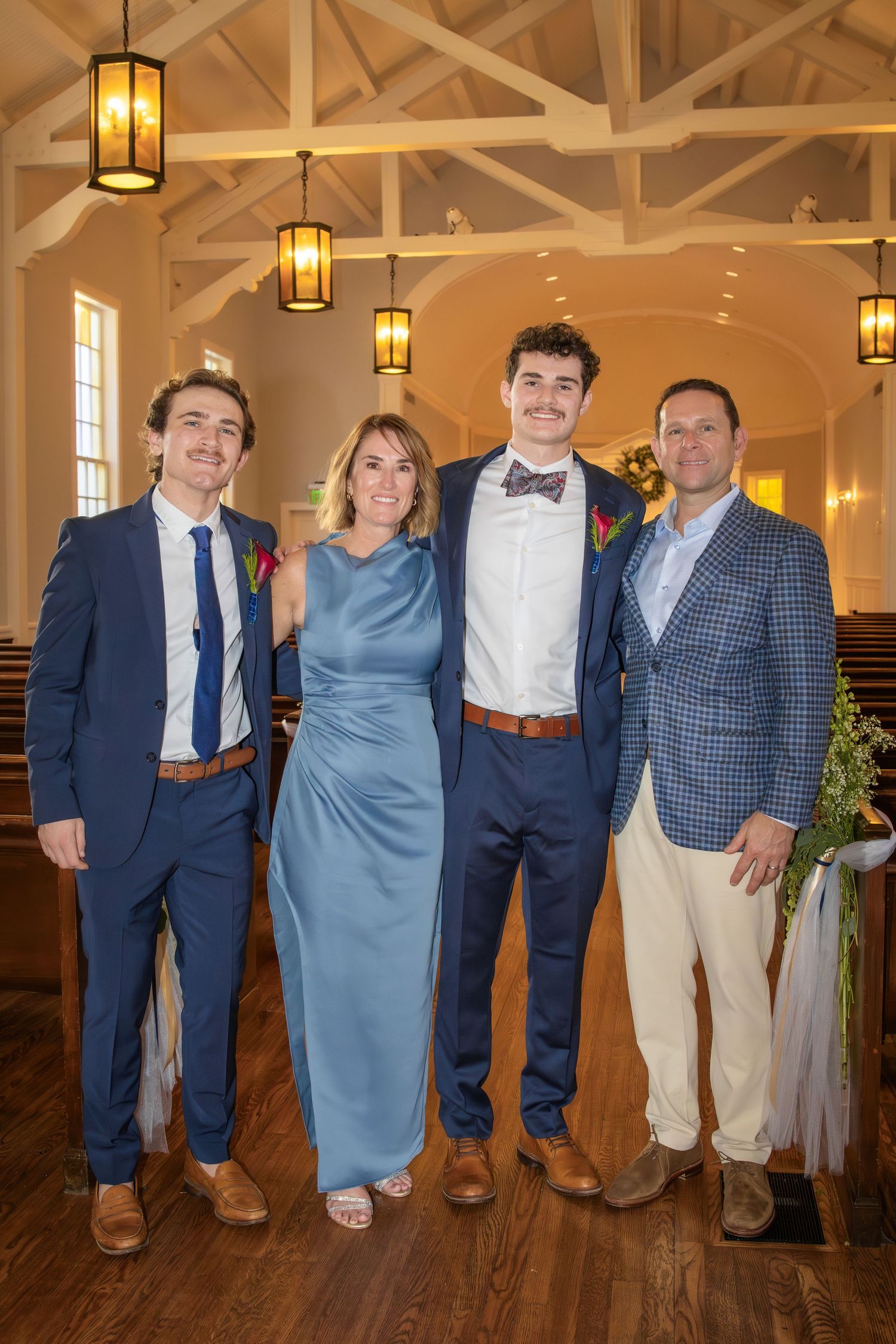 Wedding guests in blue and navy attire pose together on wooden floor under pendant lights.