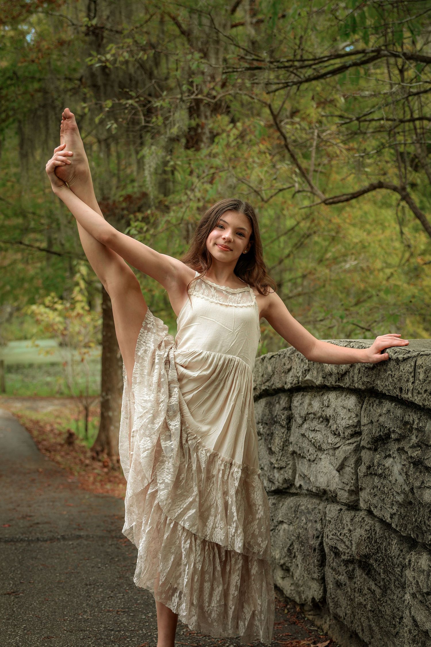 A ballet dancer in a cream lace dress performs a high leg extension against a stone wall.
