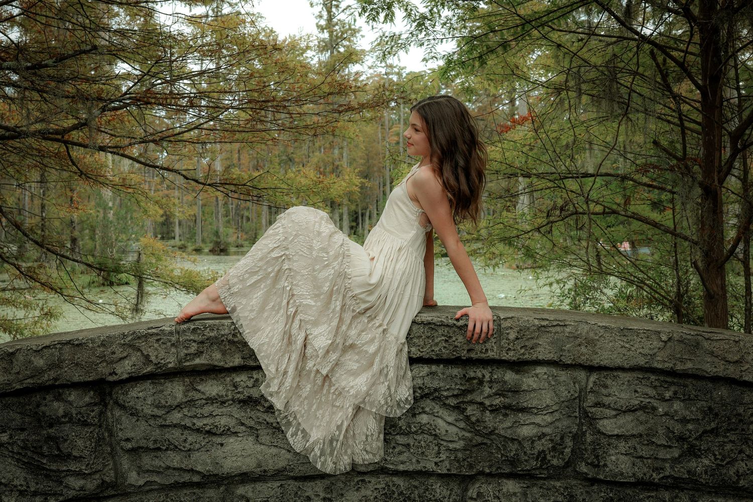 A dancer in a flowing white dress reclines on a stone wall in a dreamy forest setting.