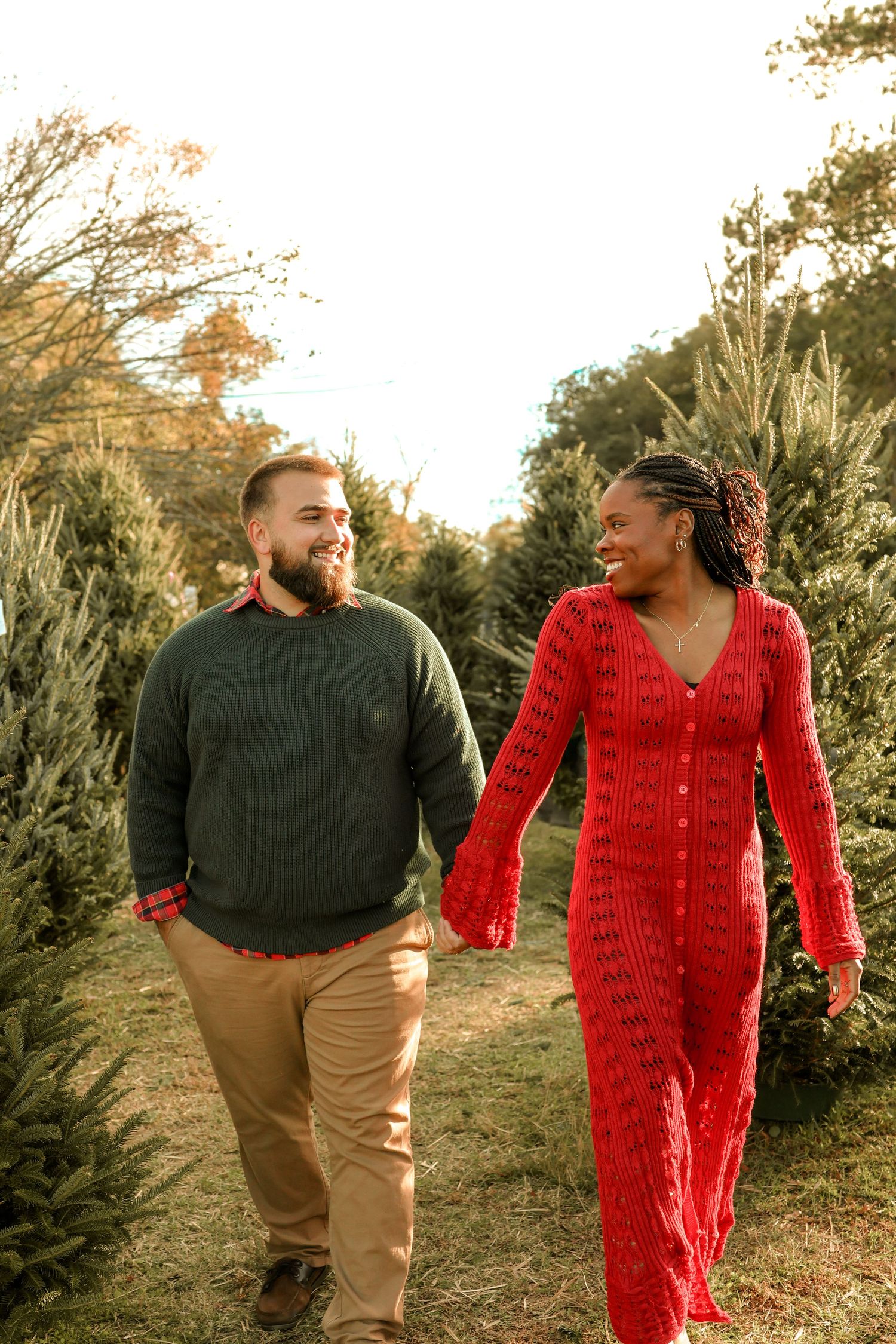 Couple in red dress and dark sweater sharing joyful moment among Christmas trees.