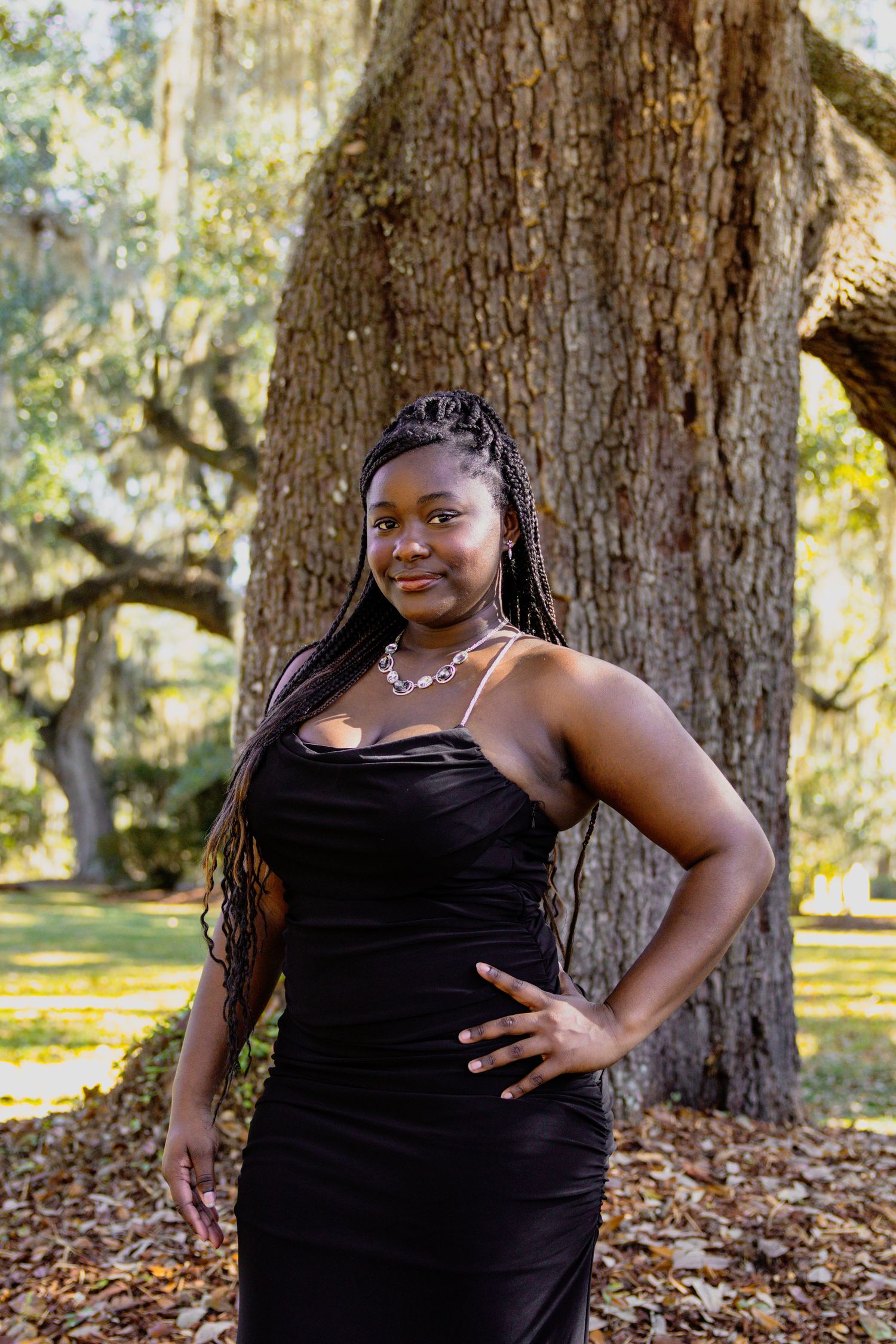A woman wearing a black formal dress poses confidently in front of a large oak tree.