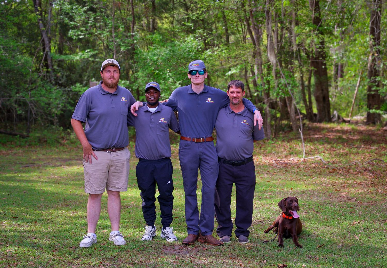 Group of workers and a black dog gathered outdoors in front of trees.