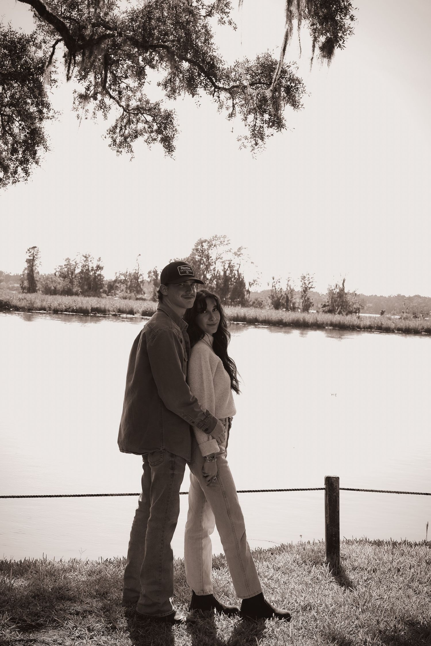 A couple embraces while standing near a wooden fence overlooking a peaceful waterfront scene.