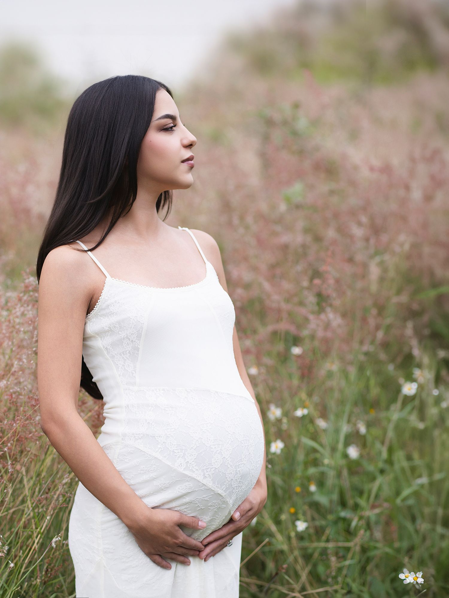 Pregnant woman in white sundress cradles her baby bump while standing in a blooming wildflower field.