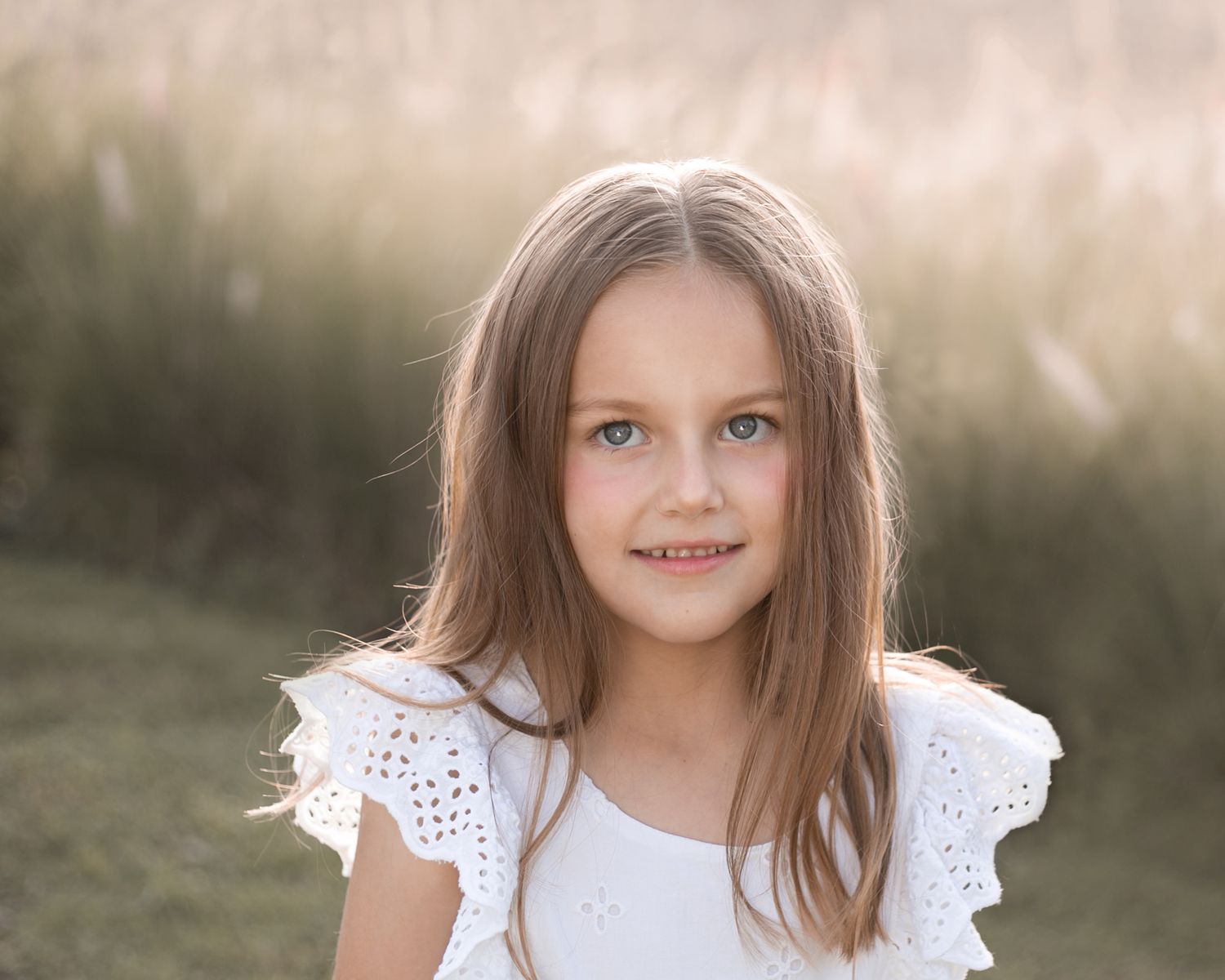 Pretty young girl with blue eyes in white ruffle dress photographed outdoors in warm golden light.