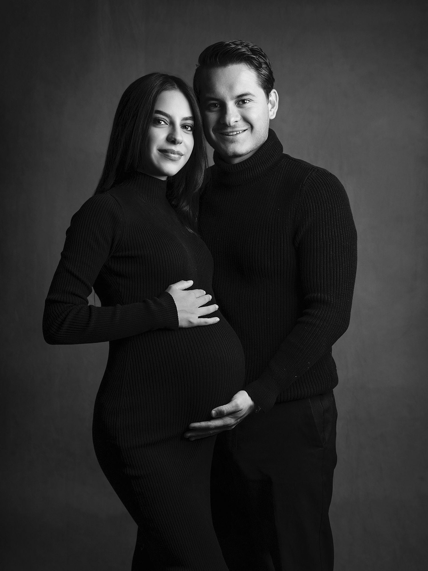 Black and white maternity photo of couple in dark outfits embracing and cradling pregnant belly in studio.