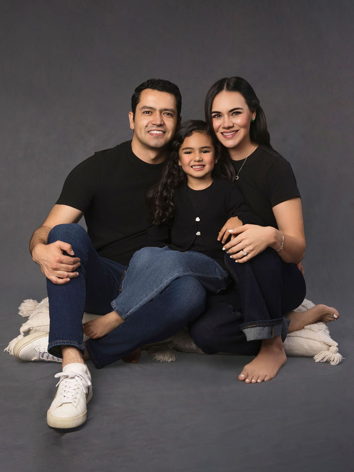 A cheerful family of three in matching black outfits posing for a studio portrait on gray background.