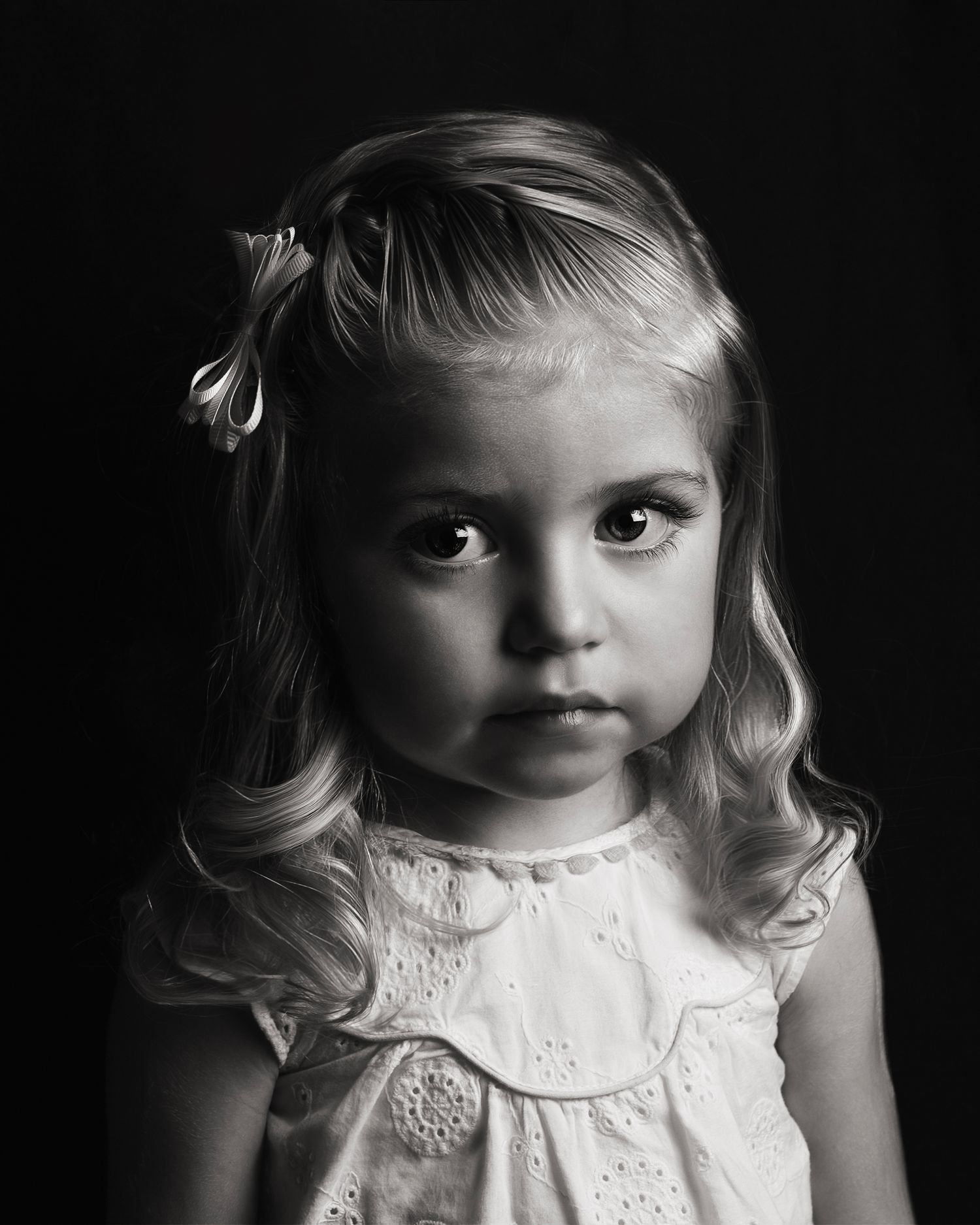 Toddler girl with blonde curls and hair bow in white dress in dramatic black and white portrait.