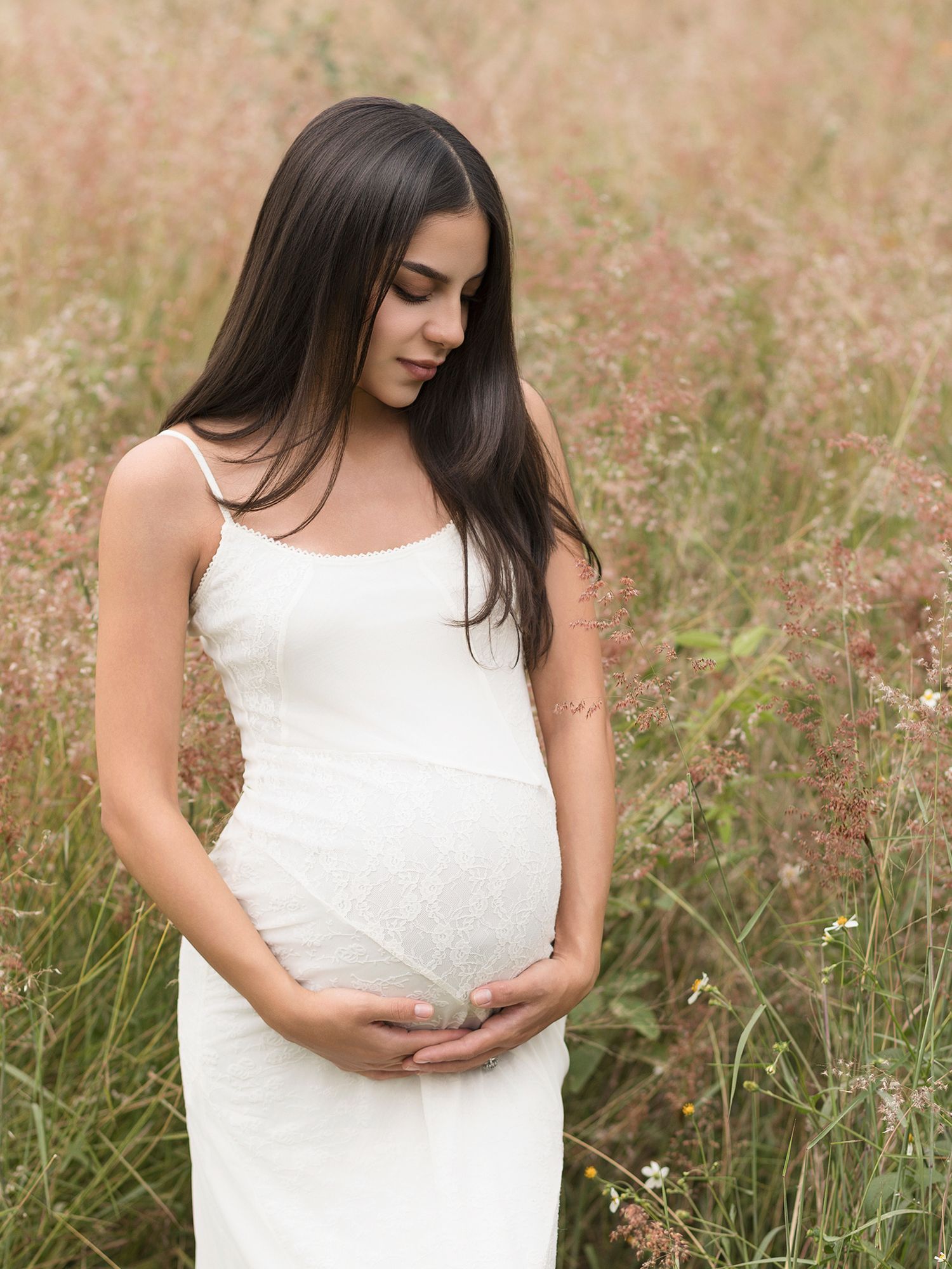 Pregnant woman in white dress holding baby bump in a field of wildflowers.