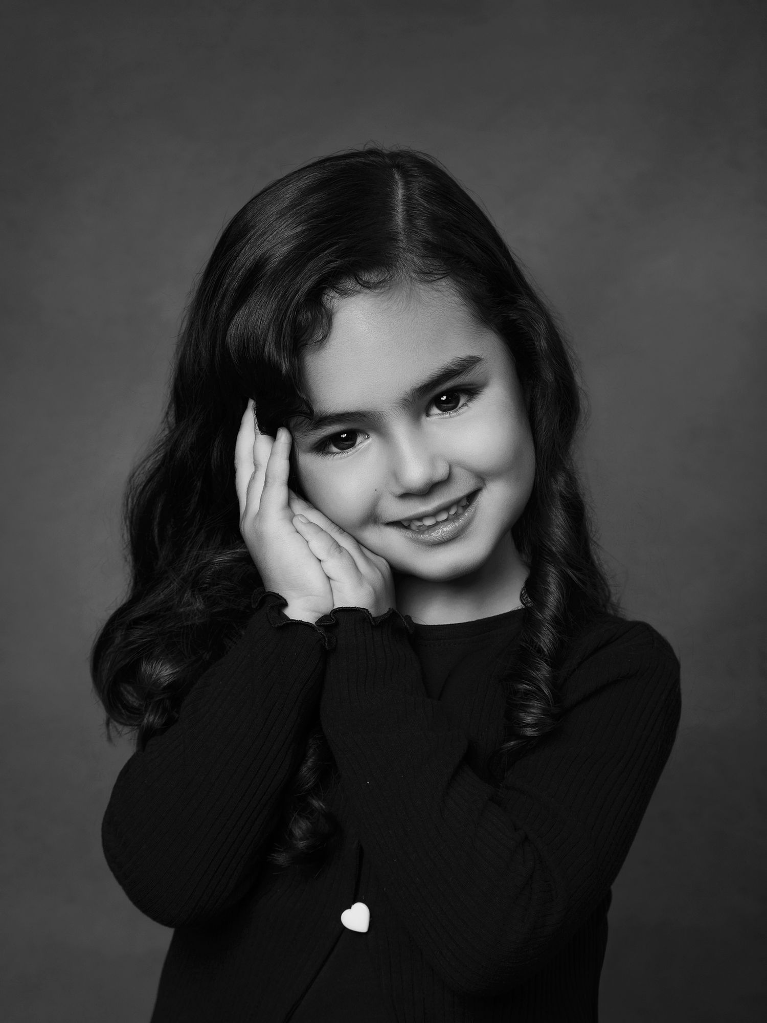 Black and white studio portrait of smiling young girl with dark wavy hair, hand on cheek.