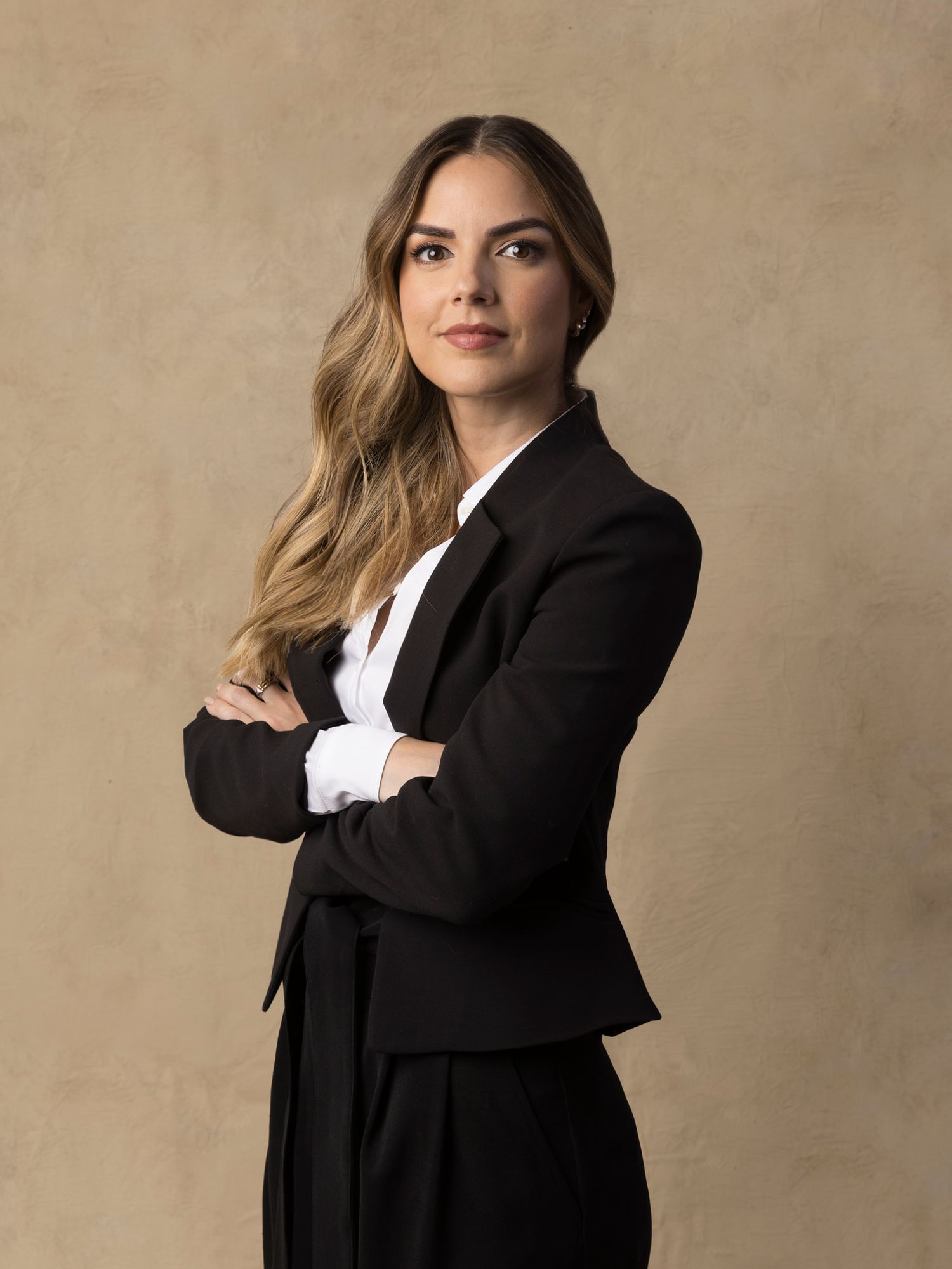 Professional woman in black blazer and white shirt poses with arms crossed in studio.