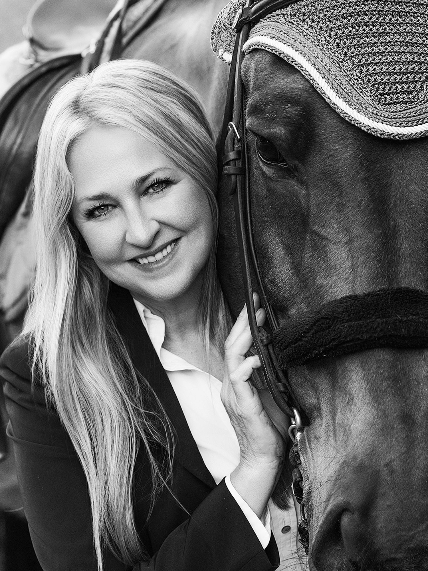 Black and white portrait of a smiling blonde woman posing closely with a dark horse wearing a bridle.