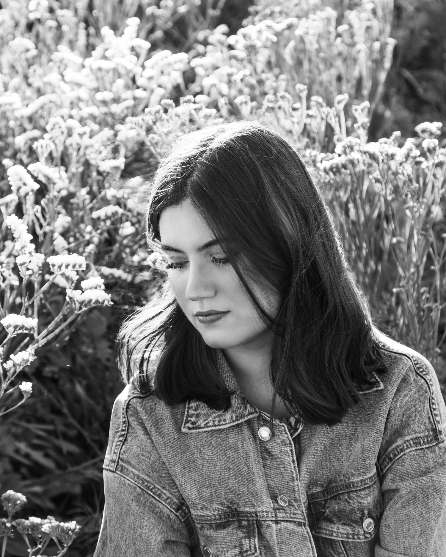 Black and white photo of a dark-haired young woman in denim jacket sitting among wildflowers outdoors.