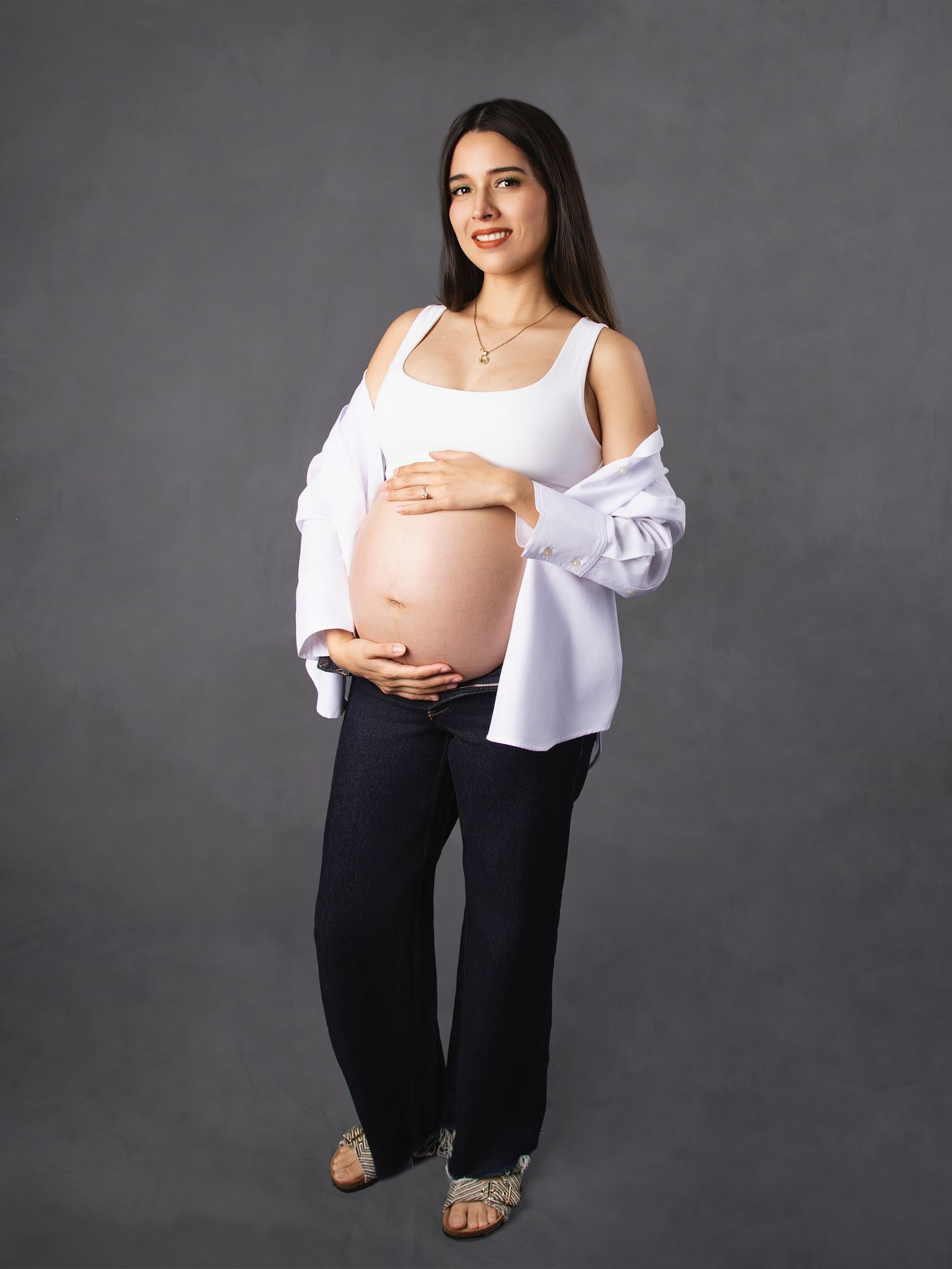 Smiling pregnant woman in white top and jeans showing baby bump in studio.