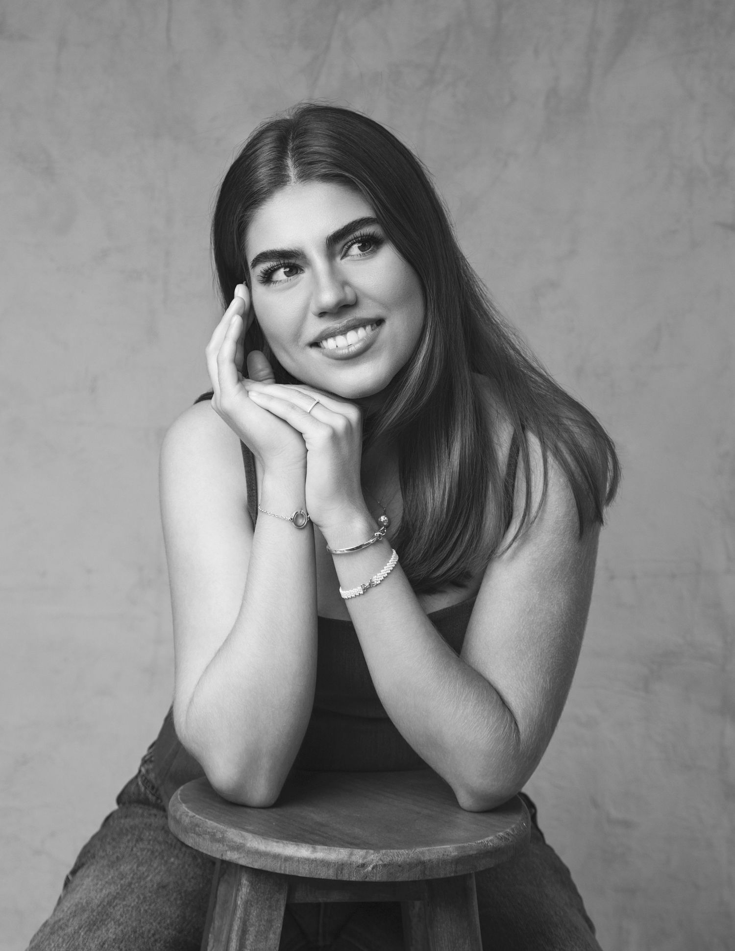 Black and white portrait of young brunette woman smiling with hands clasped under chin on stool.
