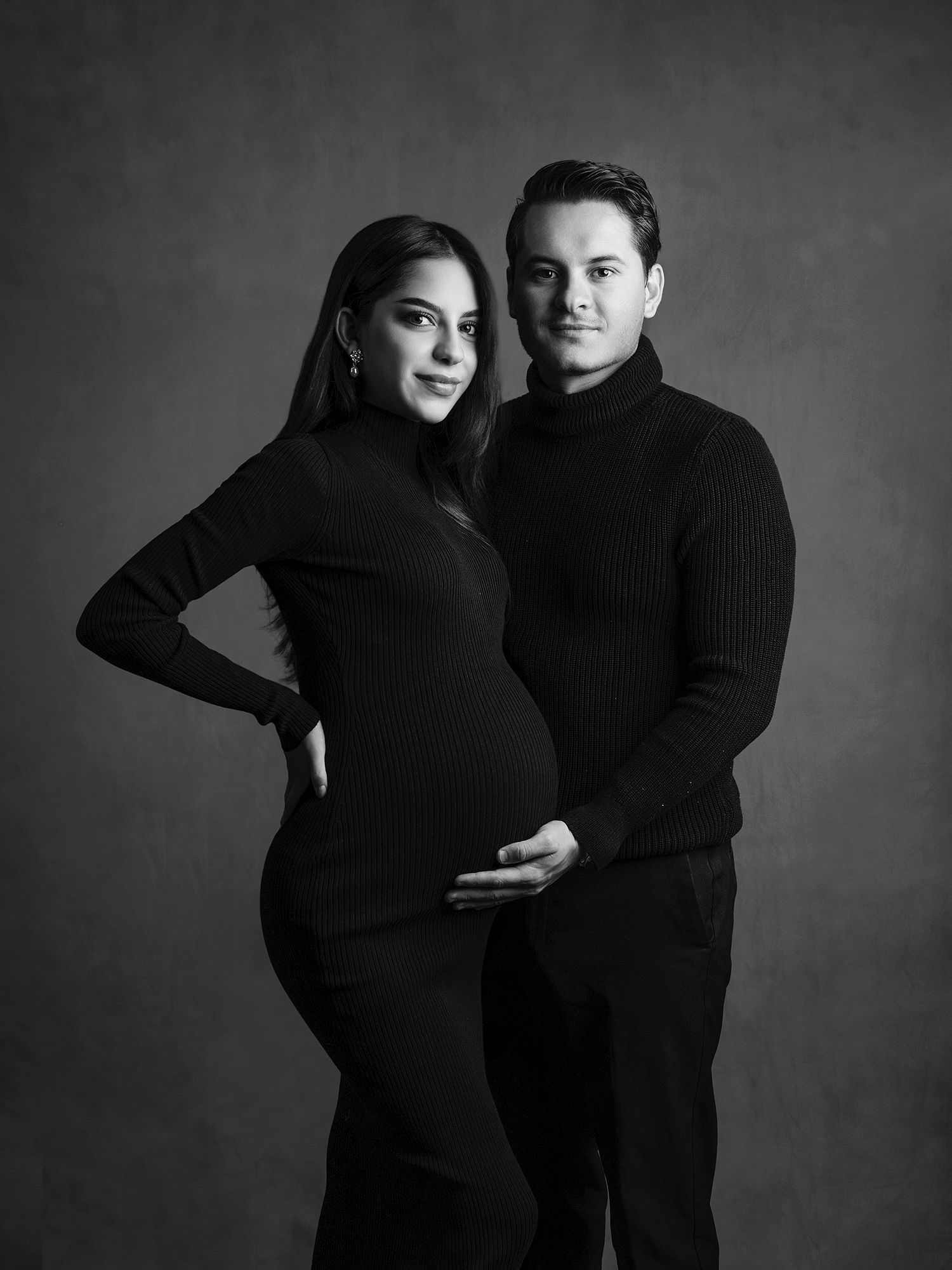 Elegant black and white studio maternity portrait of expecting couple dressed in all black against dark backdrop.