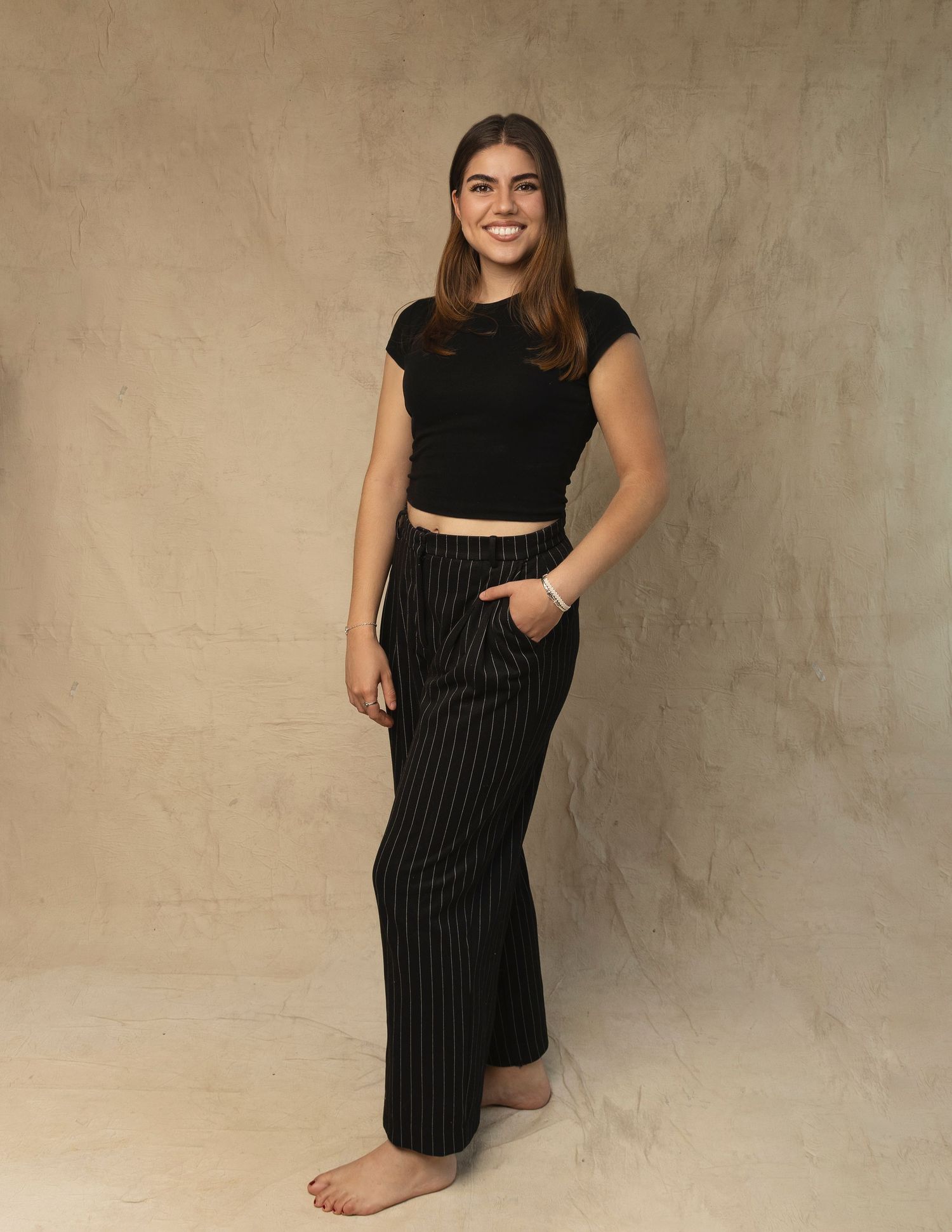 Young woman in black crop top and pinstripe wide-leg pants posing barefoot in studio setting.