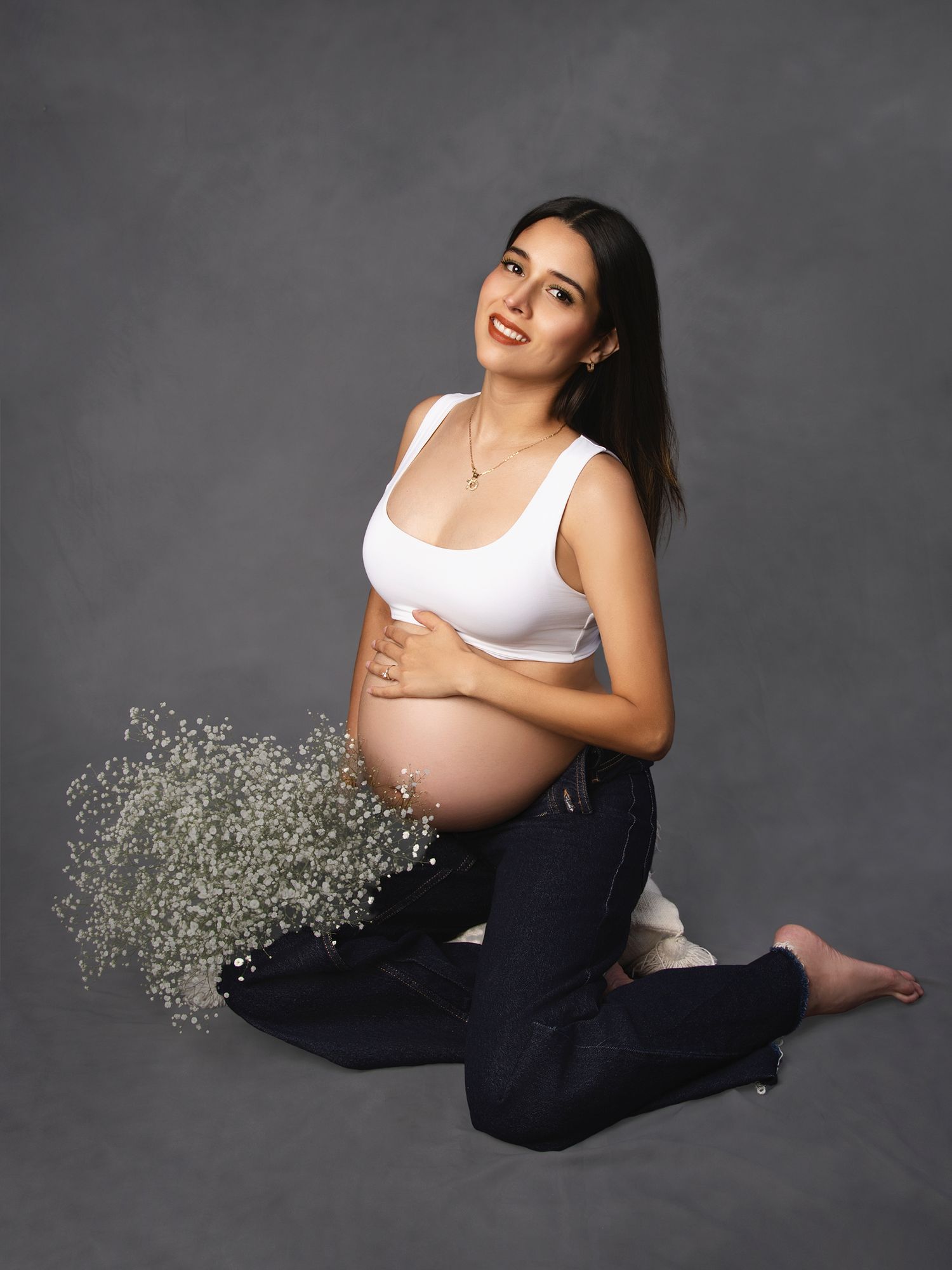 Pregnant woman in white top sitting with baby's breath flowers in studio.
