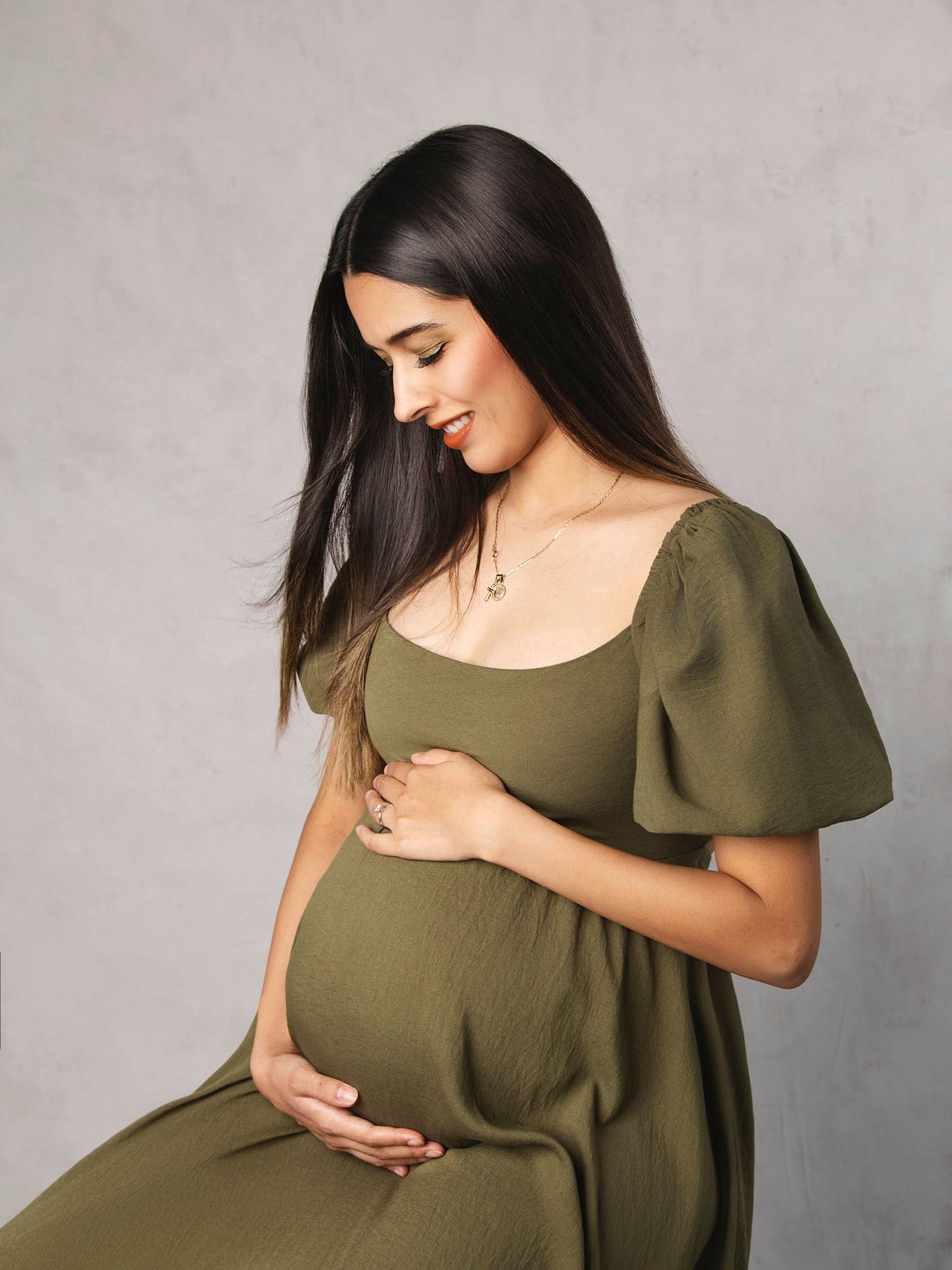 Pregnant woman in olive green dress cradling baby bump in studio.