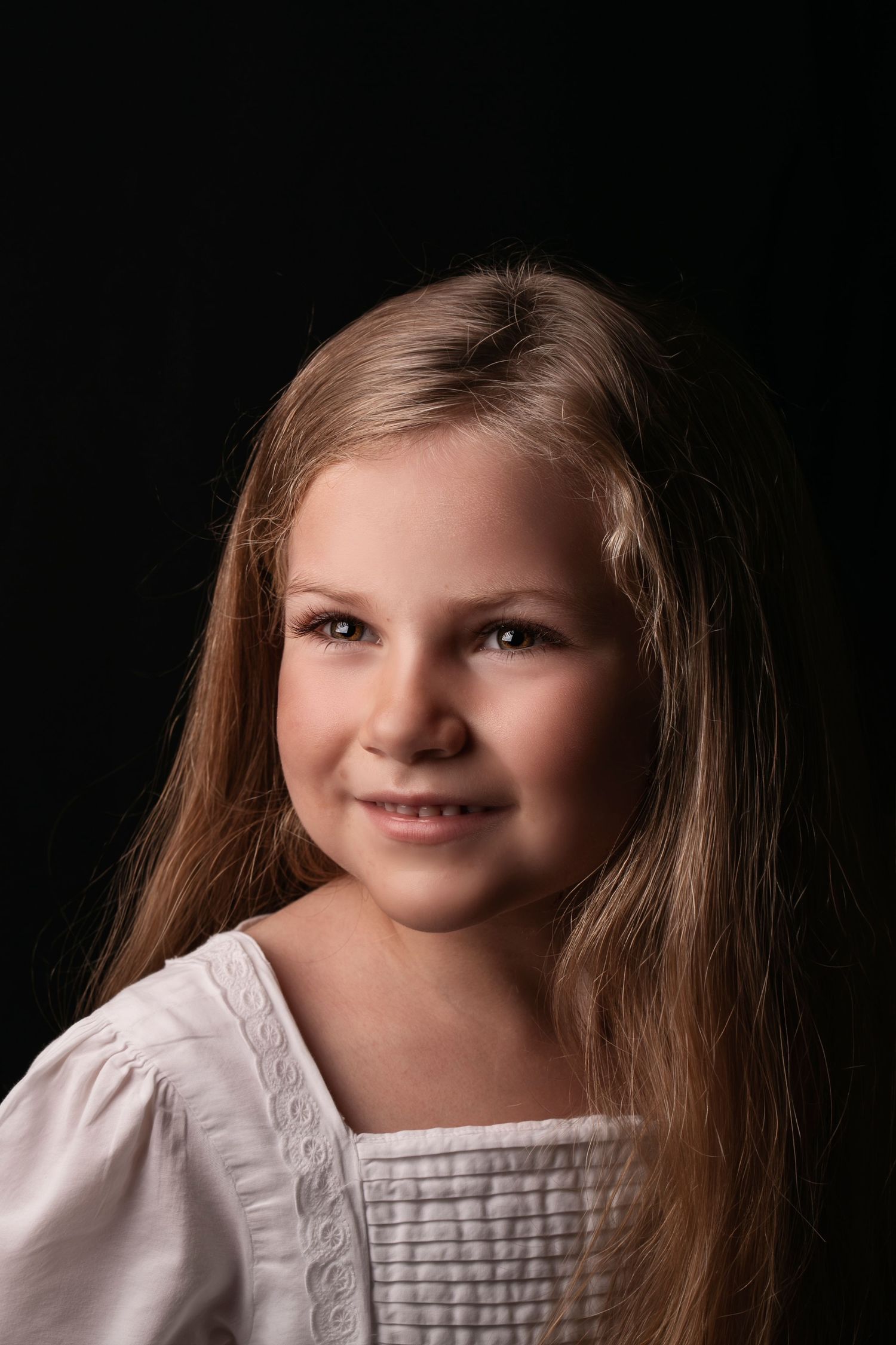 Young girl with long blonde hair smiling in a white blouse against a dark background.