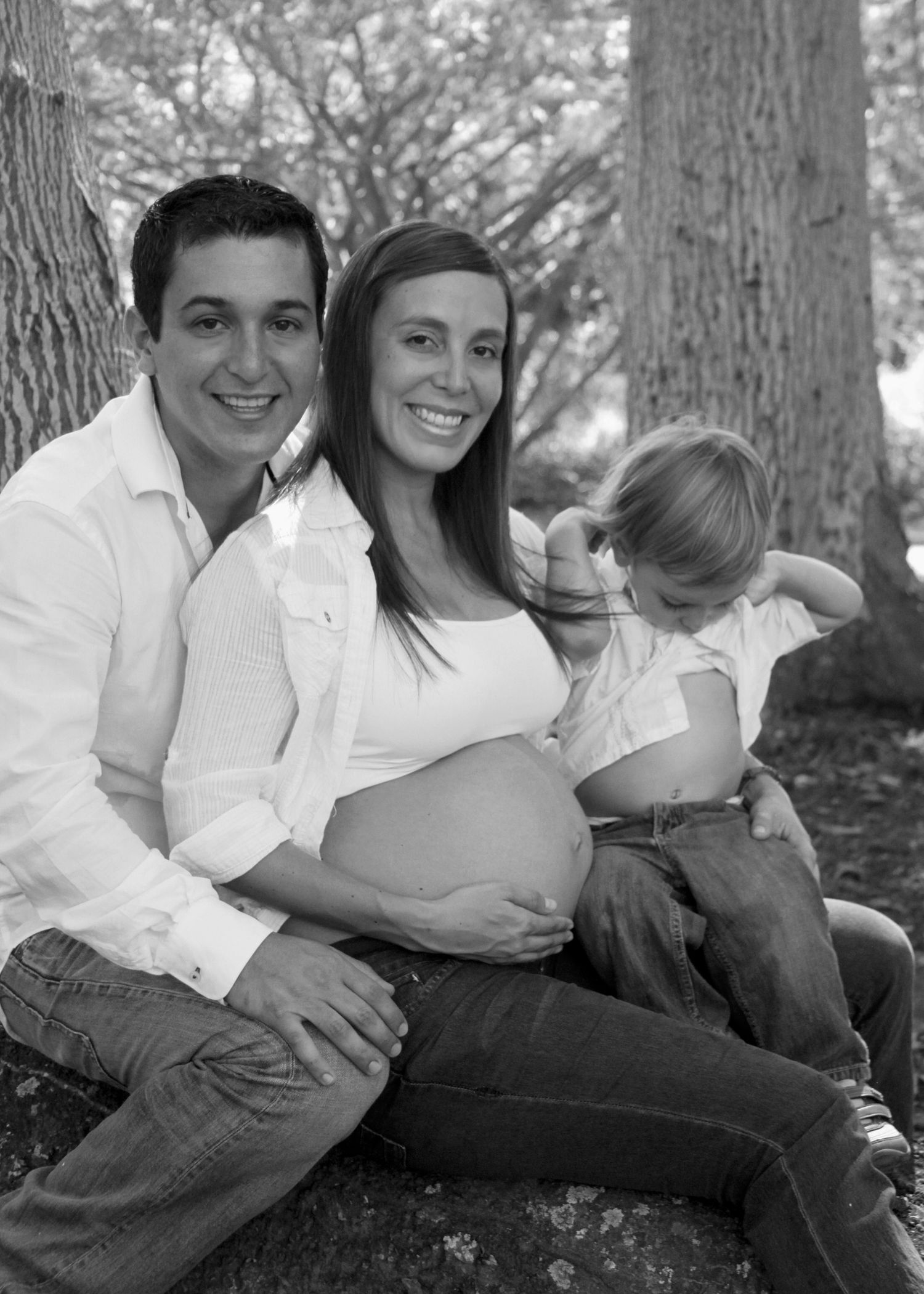 Black and white outdoor maternity photo of a couple and toddler sitting against a tree in a park.