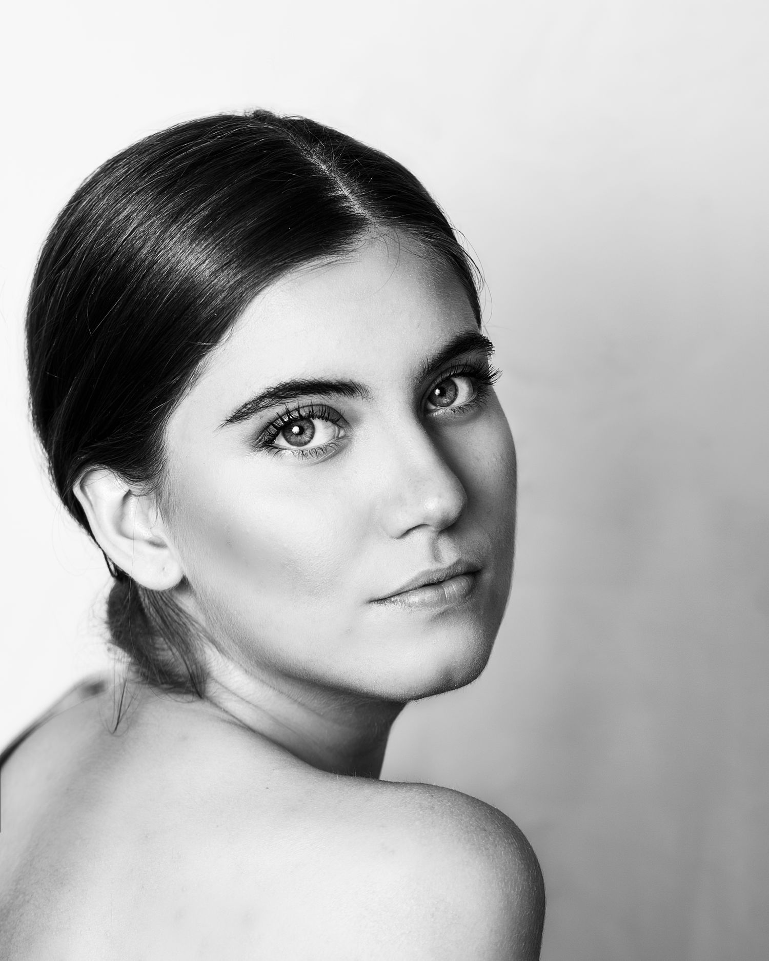 Black and white portrait of a young brunette woman with hair pulled back looking over her bare shoulder.