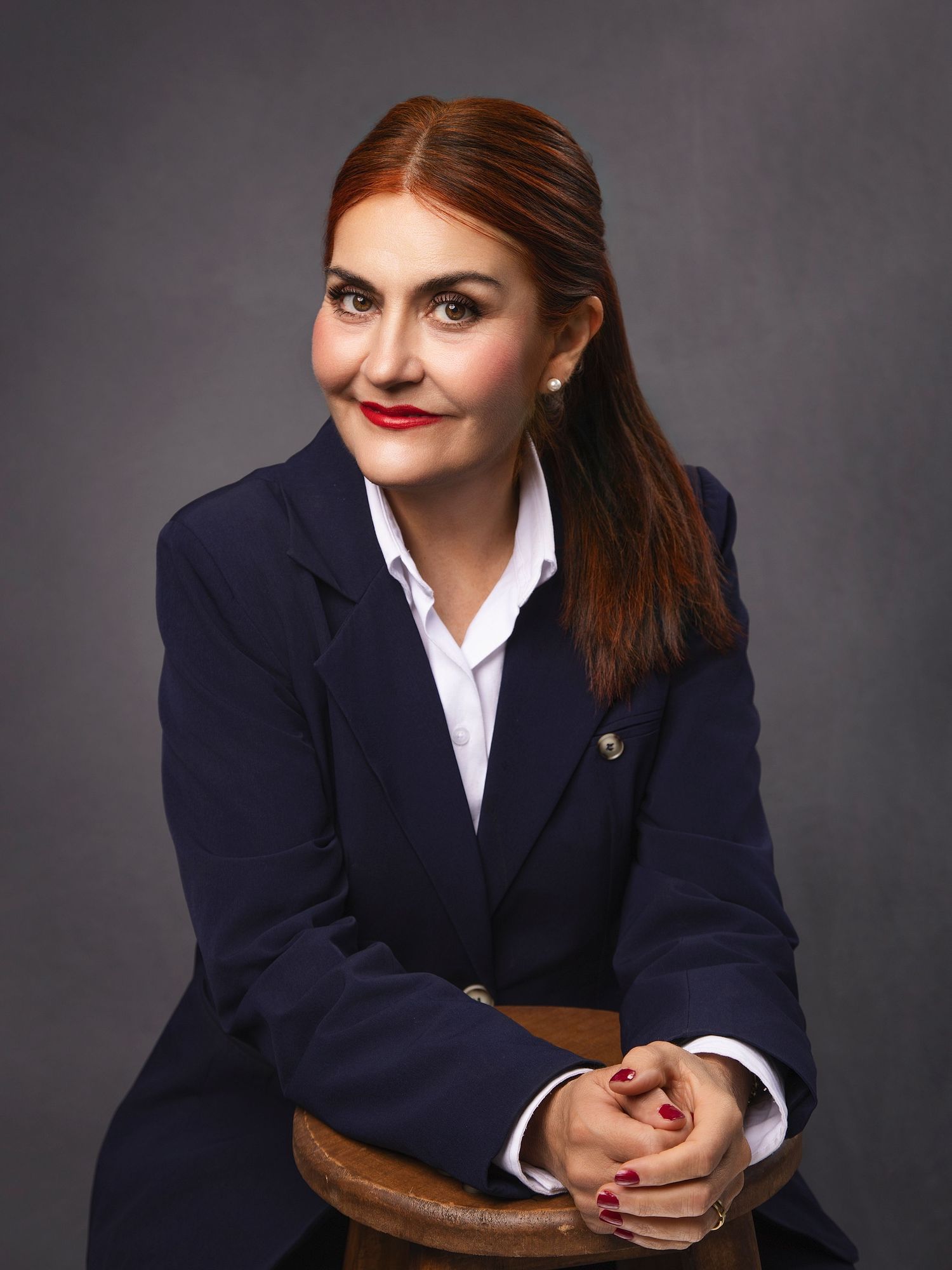 Professional woman in navy blazer with red lipstick, seated portrait against dark gray background.