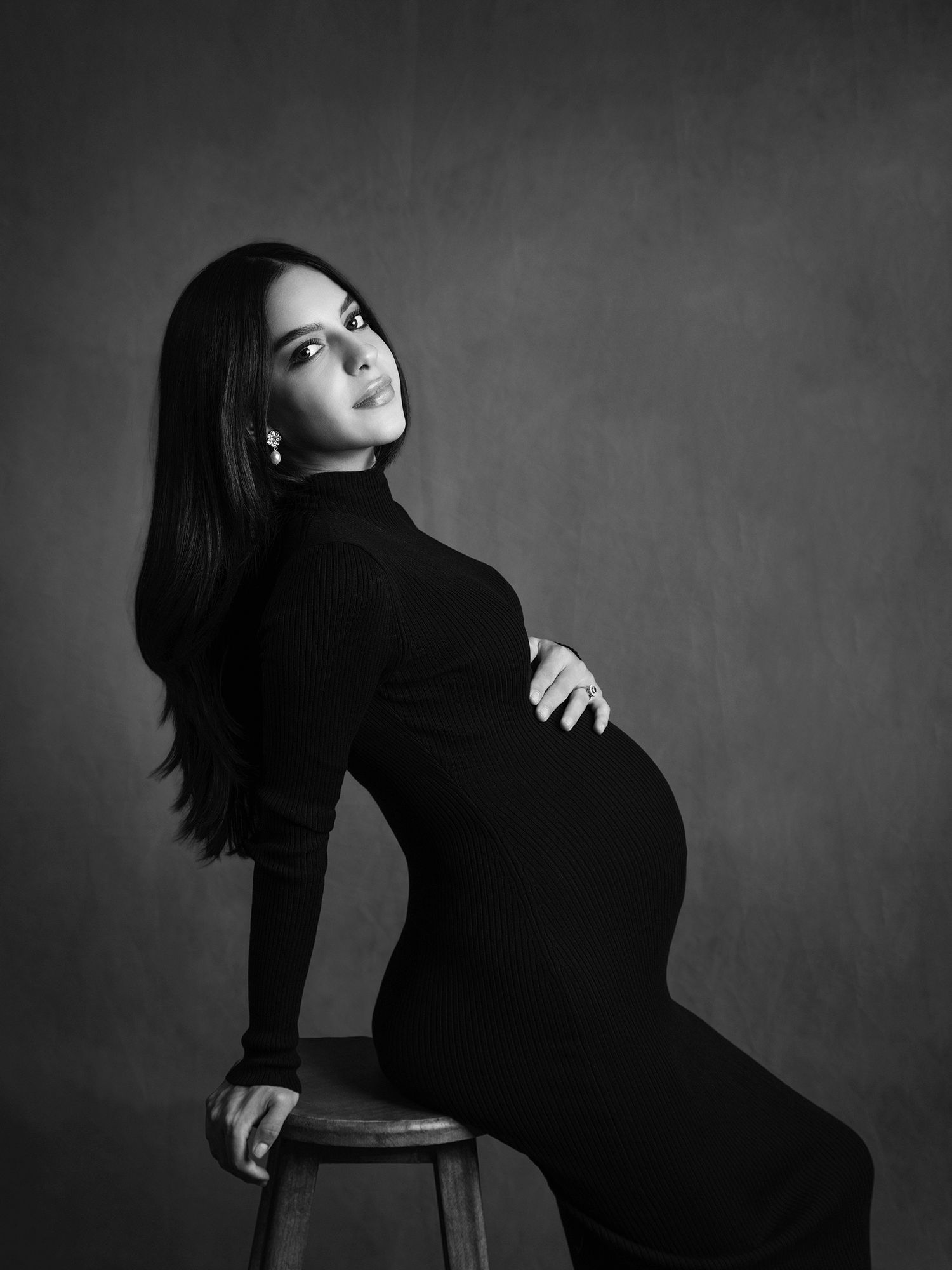 Elegant black and white studio maternity portrait of pregnant woman in turtleneck dress seated on stool.