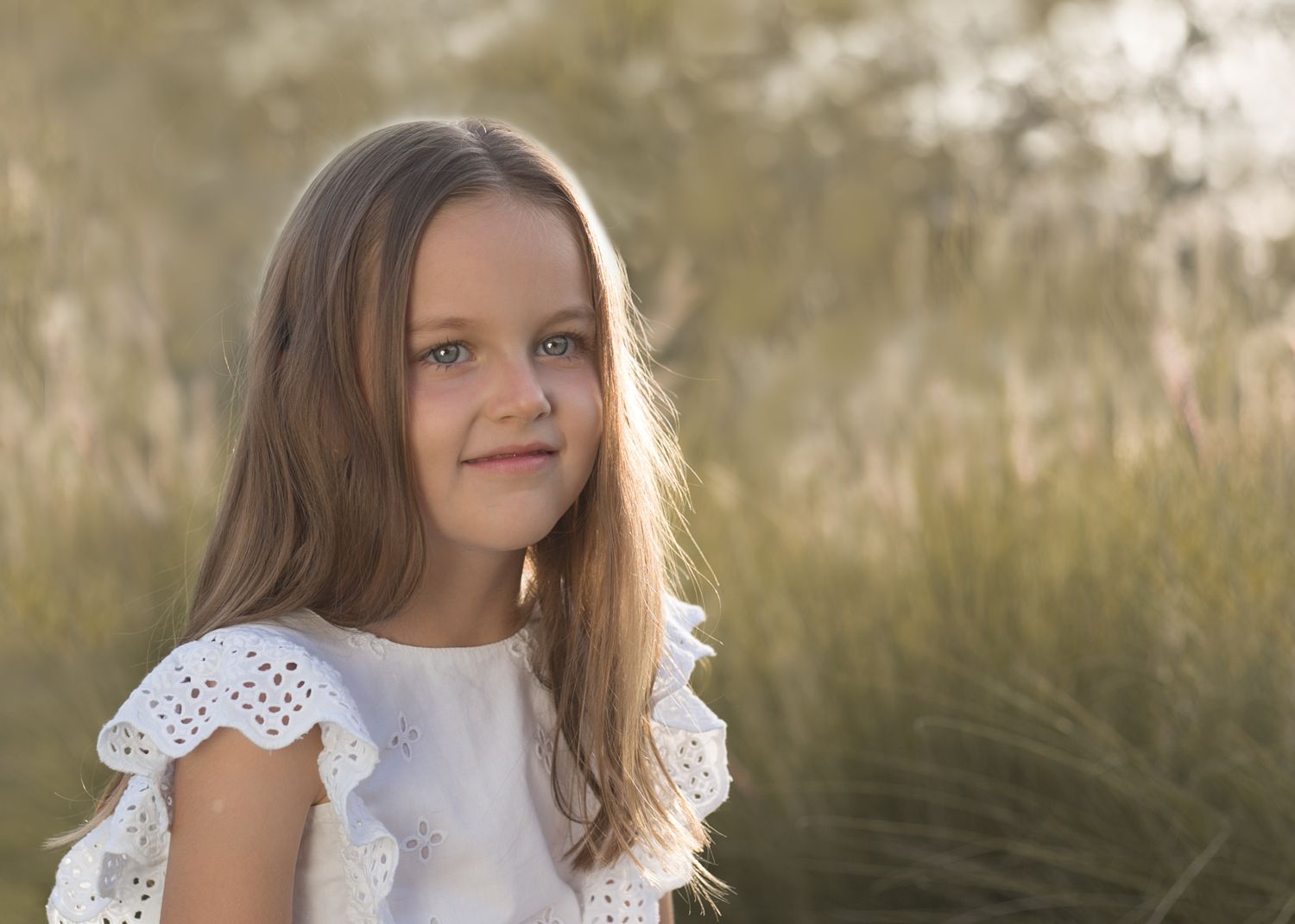 Young girl in white eyelet dress with blue eyes smiling softly in golden outdoor natural light.