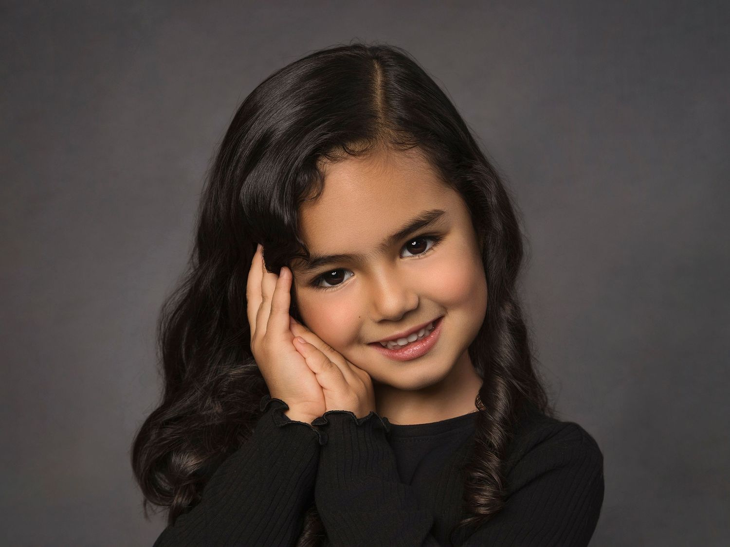 Young girl with long dark hair smiling warmly, posing with hands near face against a gray studio background.
