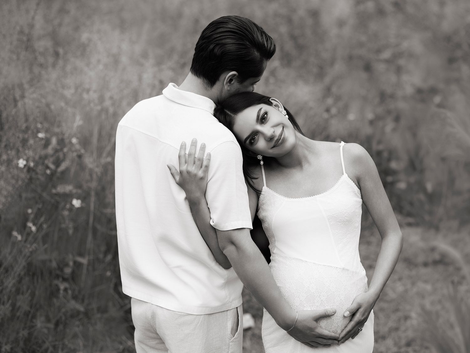 Black and white outdoor maternity photo of couple in white outfits embracing in a natural field setting.