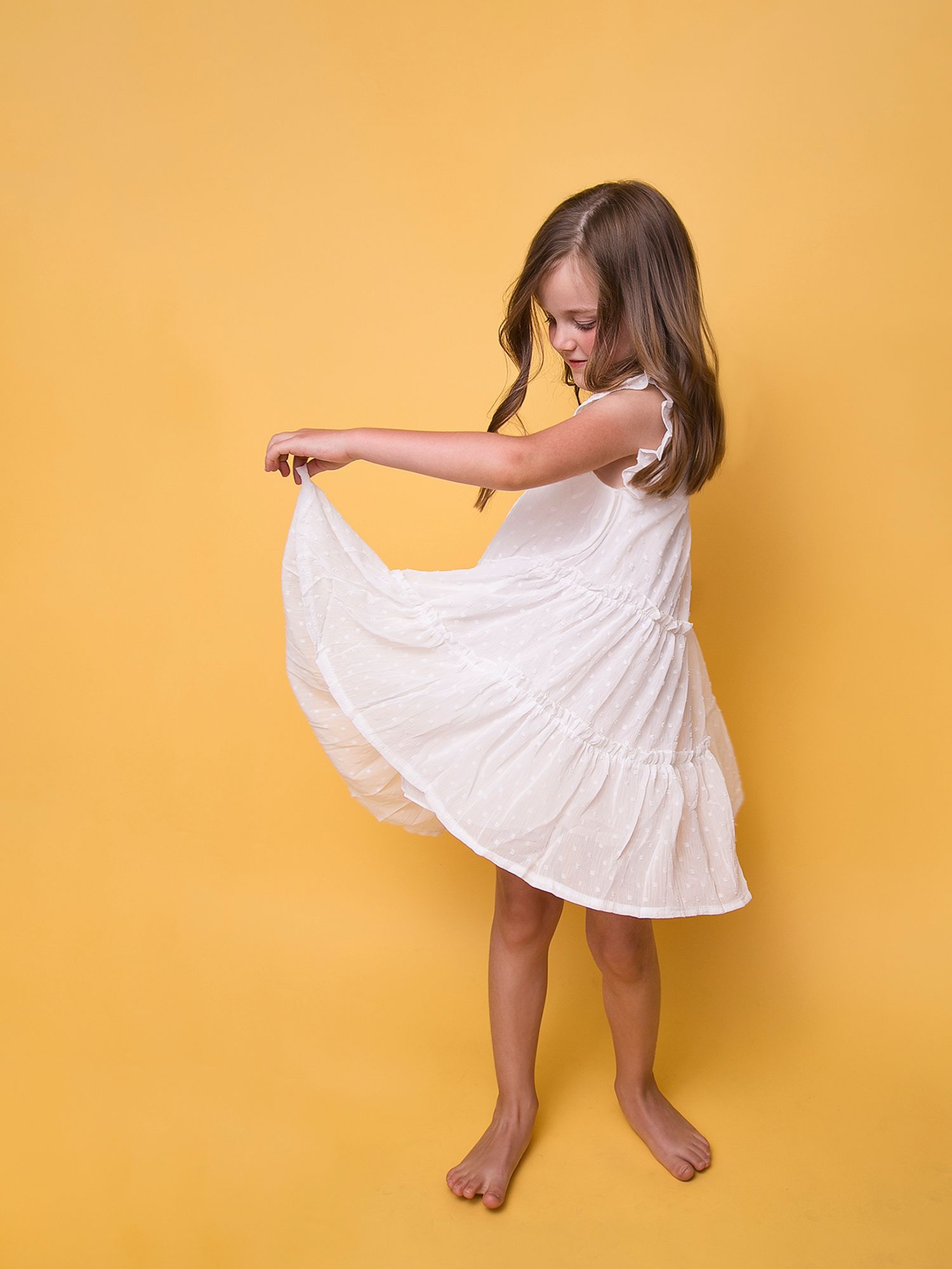 Young girl in white tiered dress twirling barefoot against bright yellow studio background.