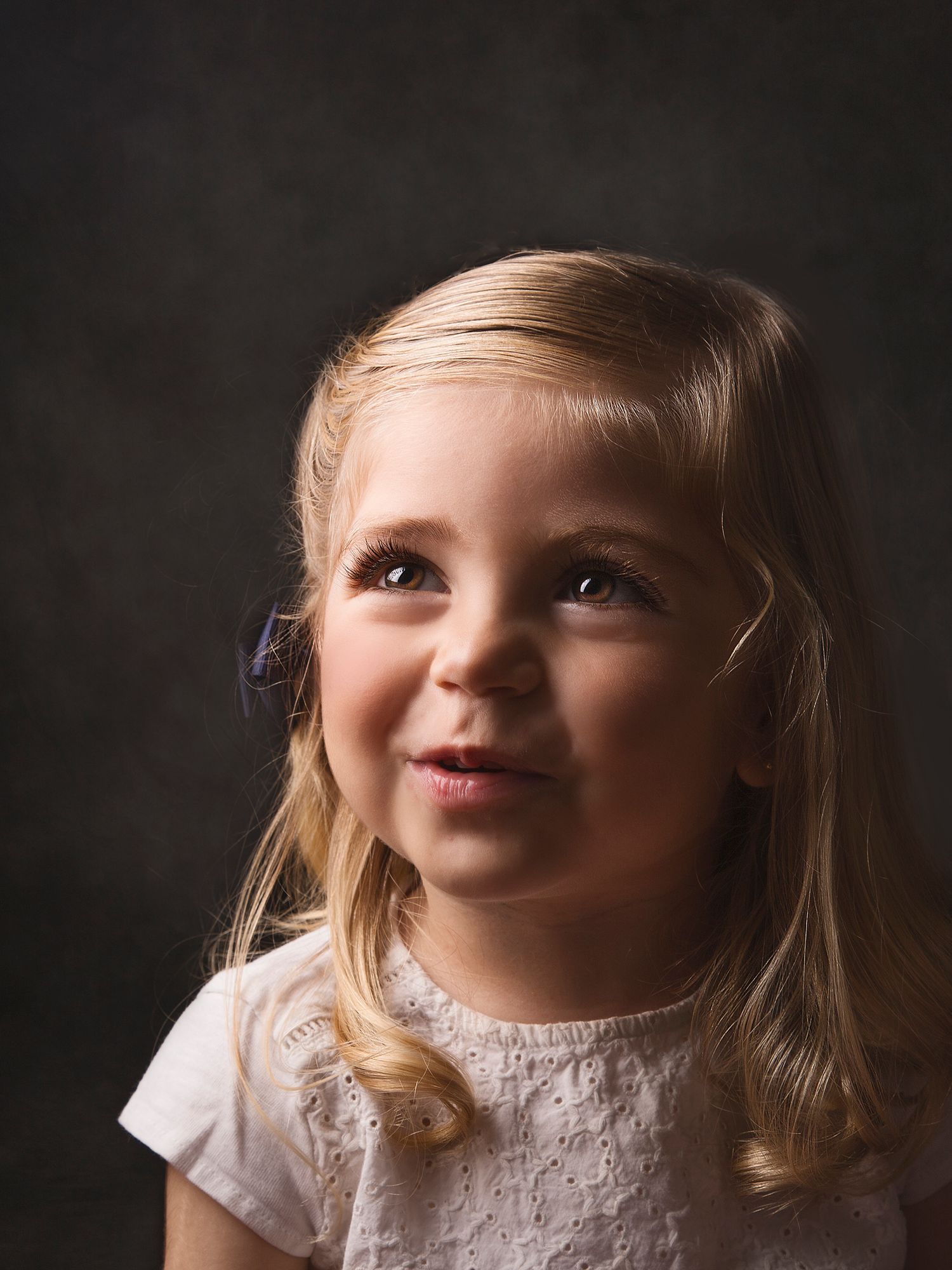Adorable blonde toddler girl in white dress looking up, dramatic studio lighting portrait.
