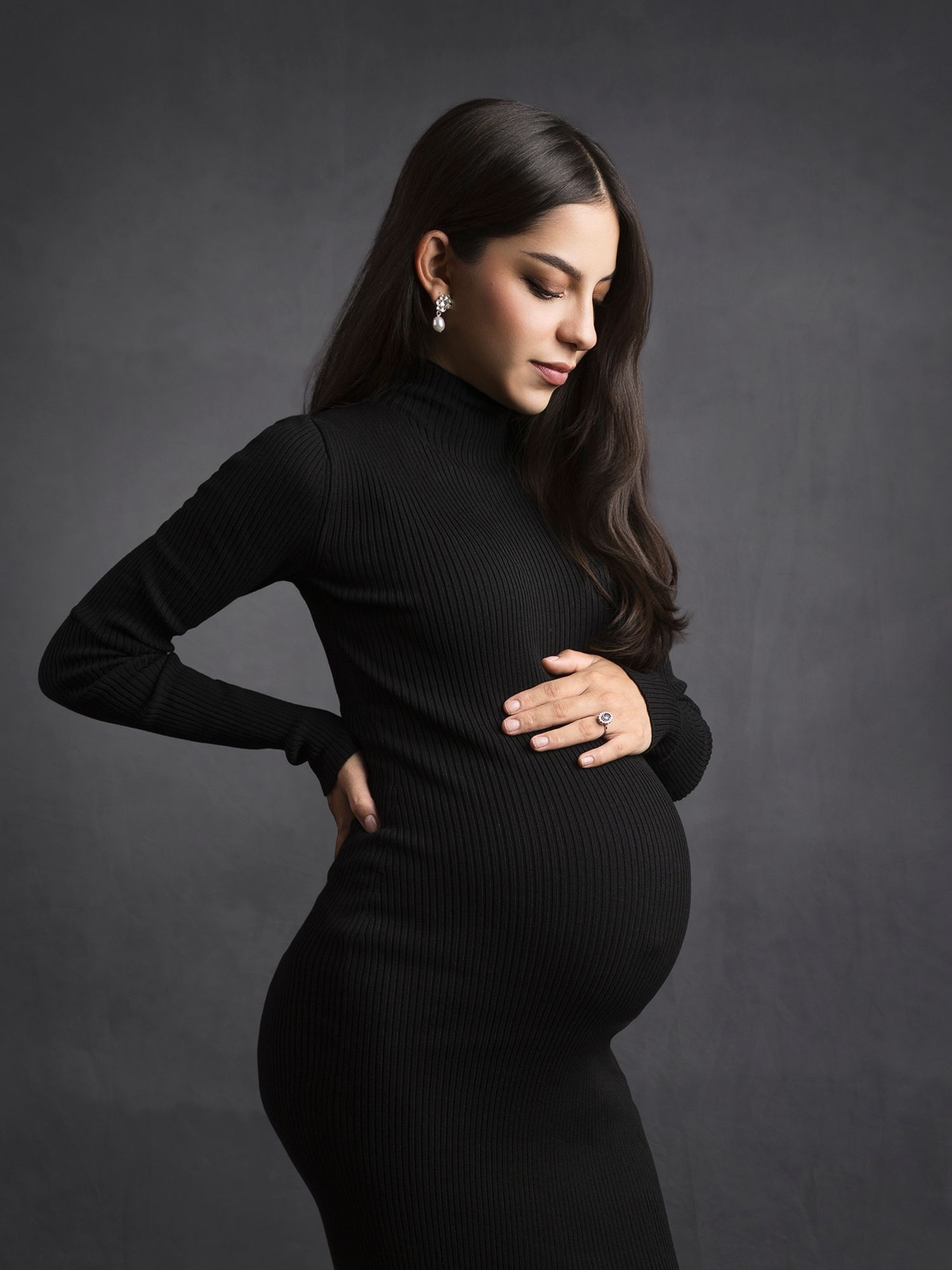 Pregnant woman in black ribbed turtleneck dress cradles her baby bump in a studio maternity portrait.