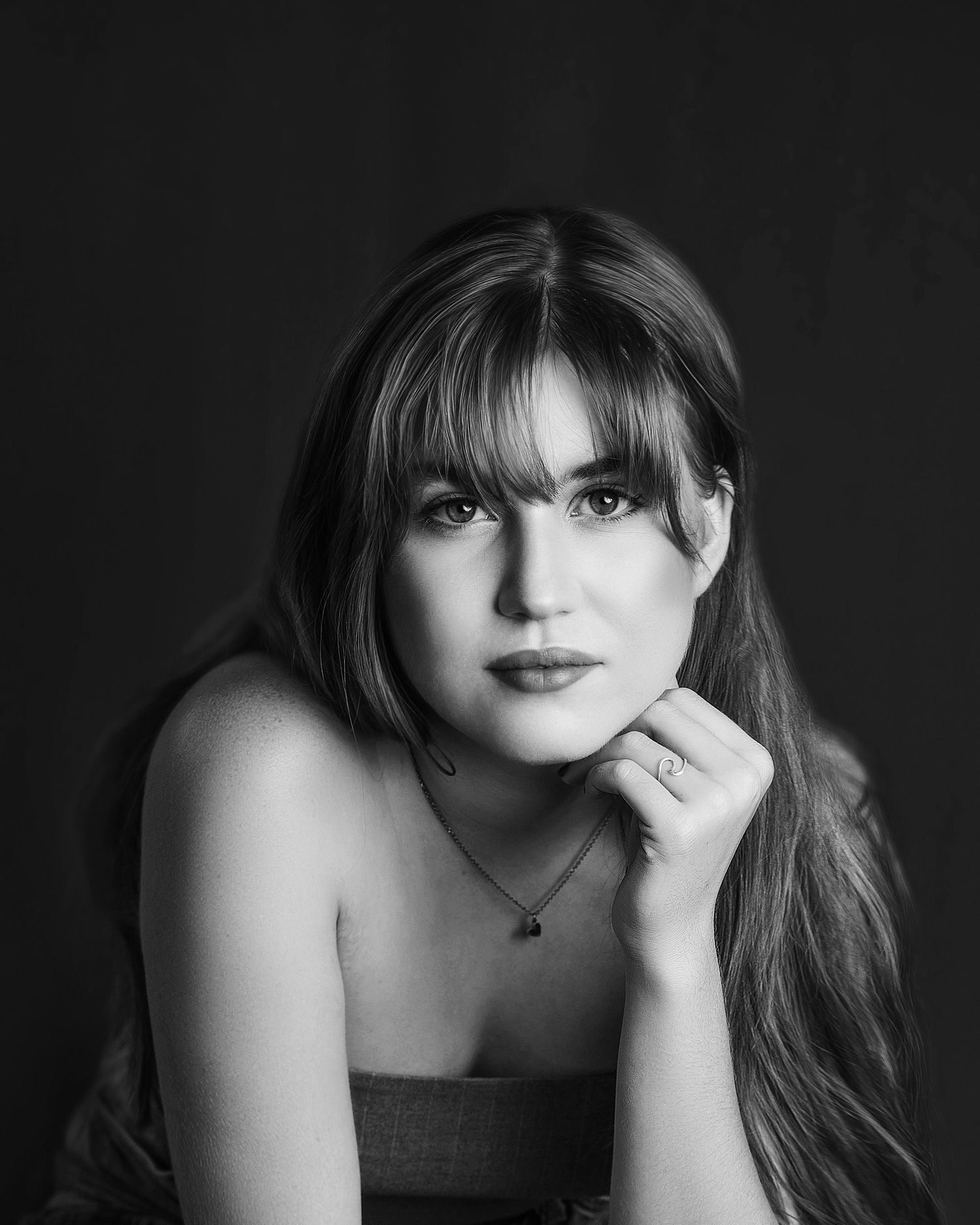 Black and white portrait of a young woman with long hair and bangs, resting chin on hand against dark background.