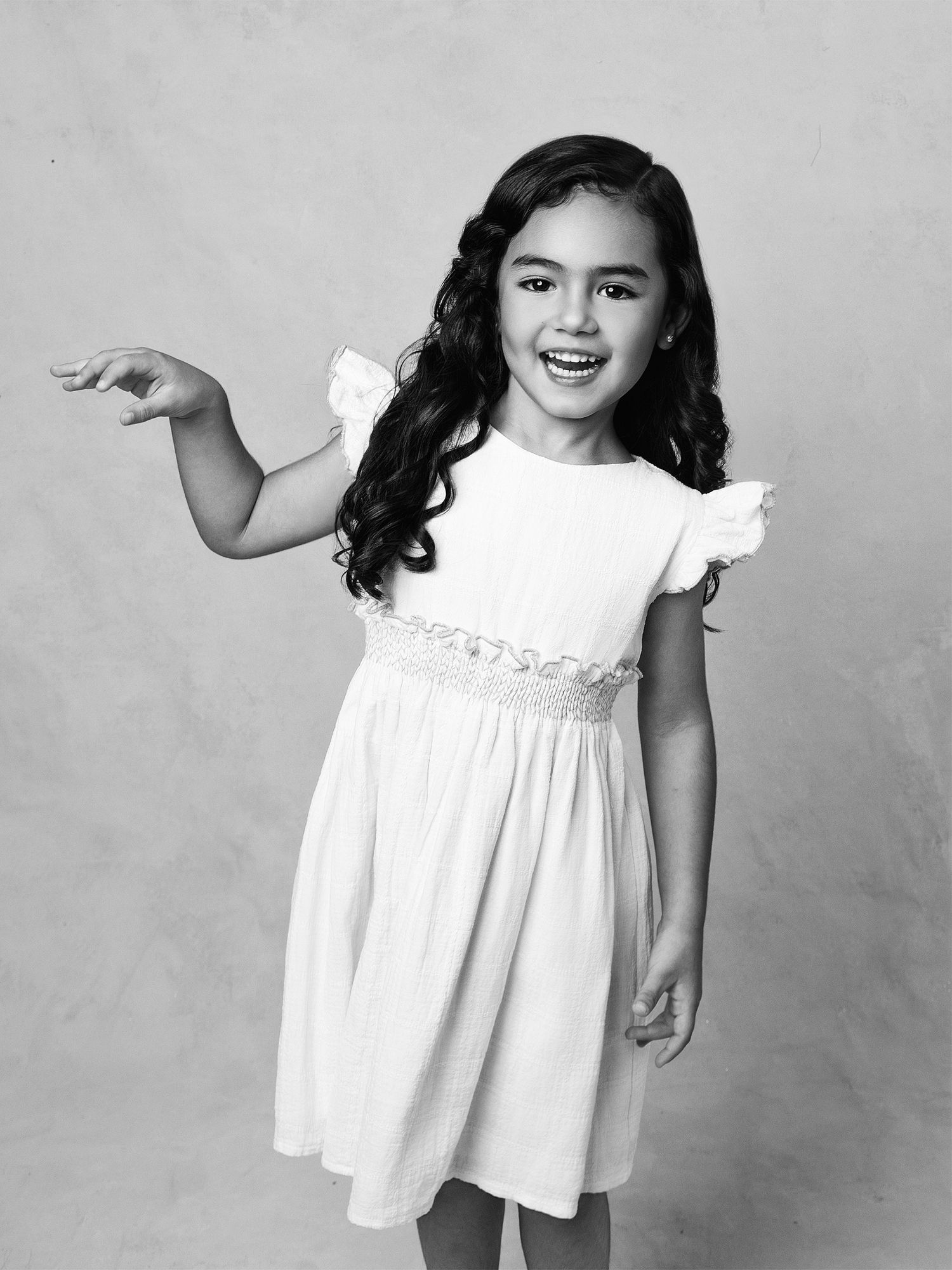 Smiling young girl in white dress with ruffle sleeves posing playfully in black and white studio portrait.