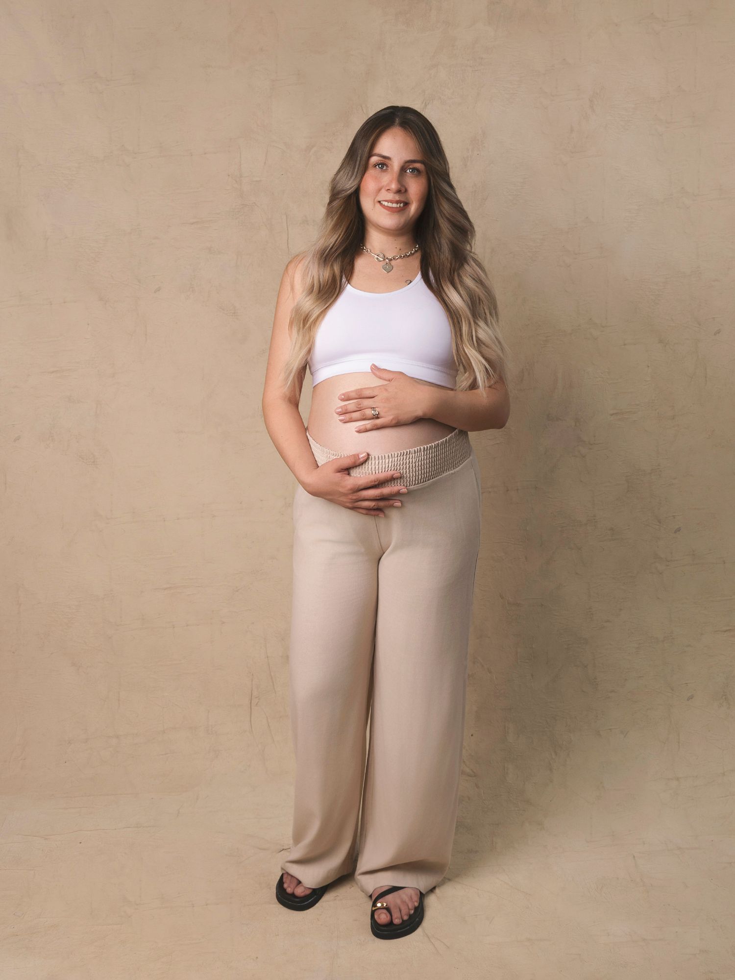 Full-length maternity portrait of a woman in white crop top and beige pants cradling her baby bump.
